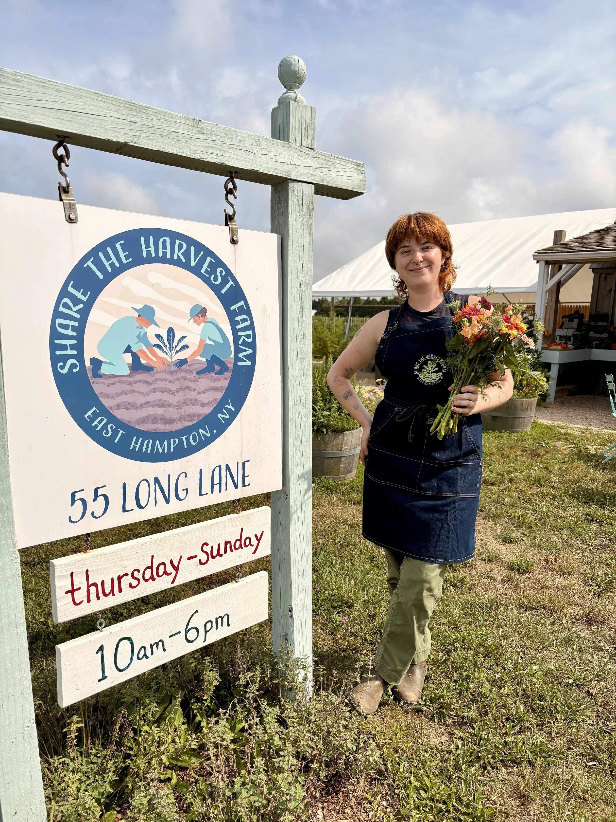 A woman holding a bouquet of flowers standing outdoors next to a sign for a farm. The sign reads 'Share the Harvest Farm, East Hampton, NY, 55 Long Lane, Thursday-Sunday, 10am-6pm.' The background includes a white tent, a small building, and green grass.