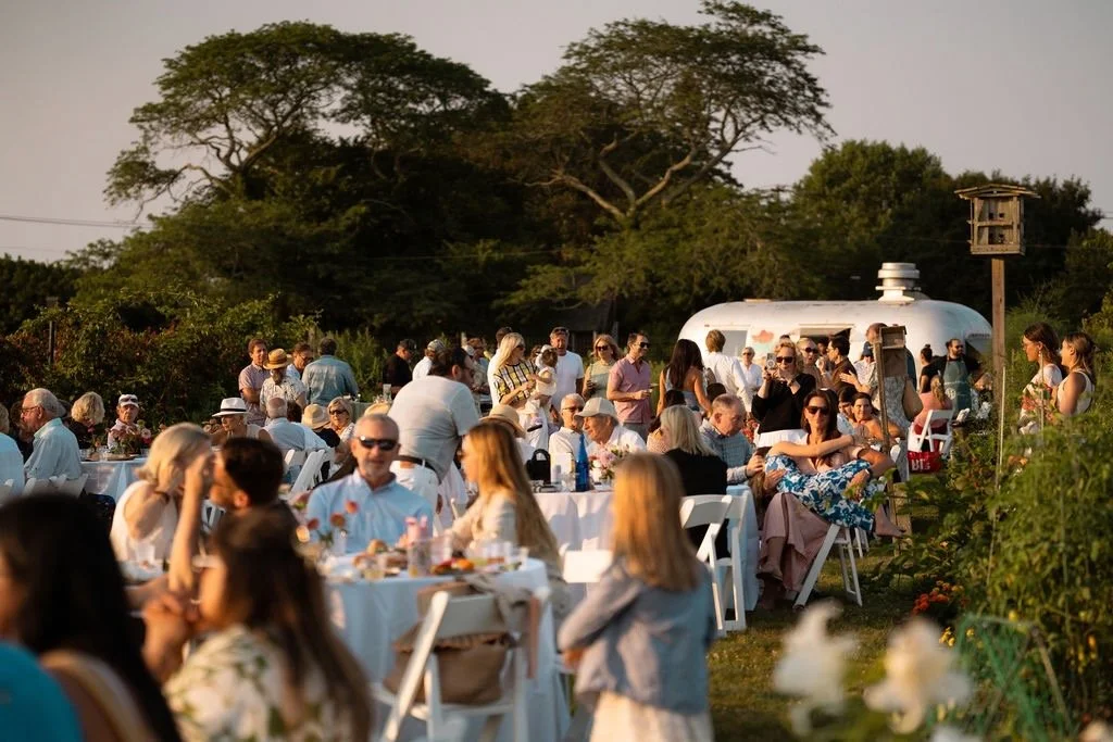 An outdoor gathering with many people seated and standing around tables, enjoying a social event or party during sunset in a garden setting with trees and a white trailer in the background.