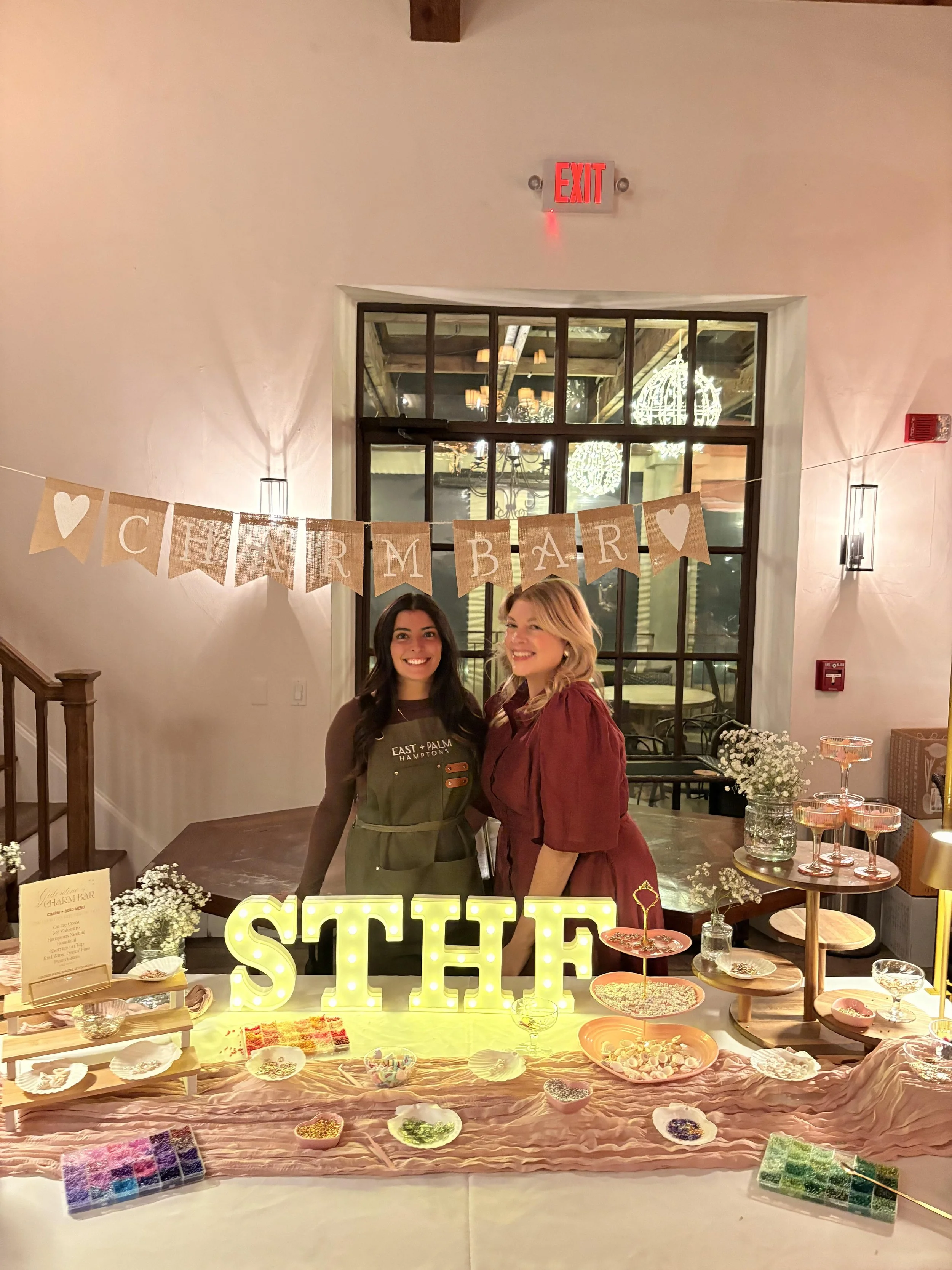 Two women standing behind a table filled with various baked goods and decorations at a party or event. A sign with lit letters spells out 'SHE'. A banner with burlap flags and white hearts reads 'CHARM BAR'.
