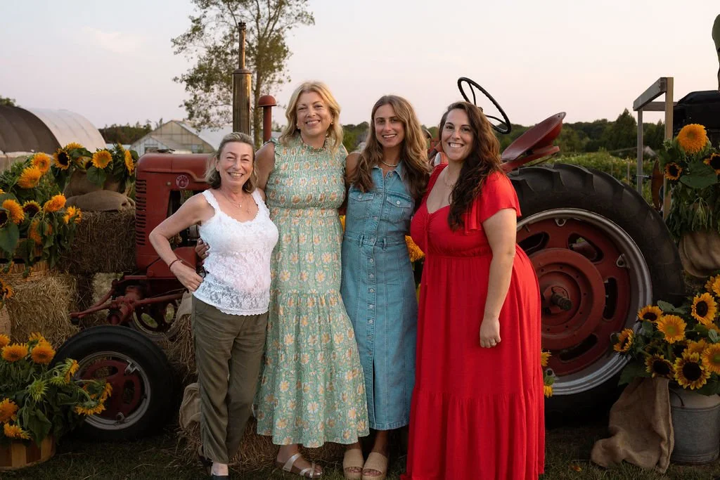 Four women standing outdoors near a vintage red tractor, surrounded by sunflowers, during sunset.