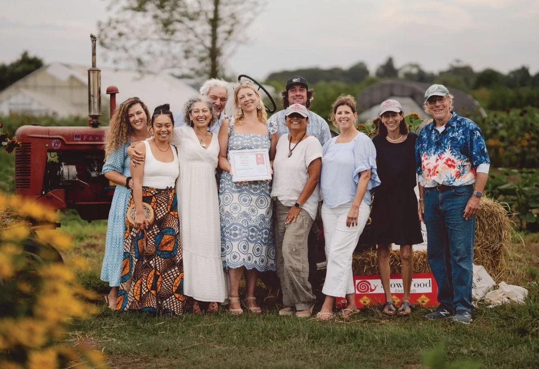Group of ten people standing together outdoors on a farm, smiling, with an old tractor, hay bales, and green fields in the background.