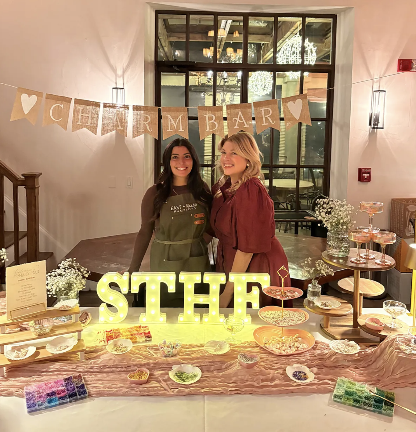 Two women standing behind a table decorated for a celebration, with a banner above them reading 'CHARM BAR' and illuminated letters spelling 'SHE' on the table. The table is filled with various colorful sweets and treats, and there are flower arrangements on either side. A large window behind reveals an indoor setting with chandeliers.
