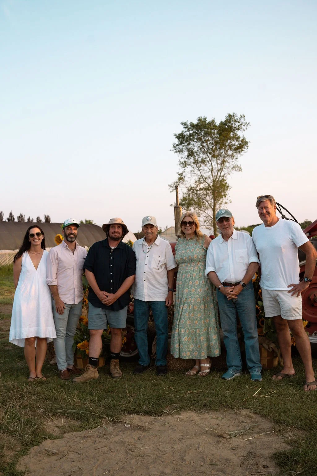 Group of eight people standing outdoors on a farm, smiling, with a tree and farm structures in the background, during what appears to be late afternoon or early evening.