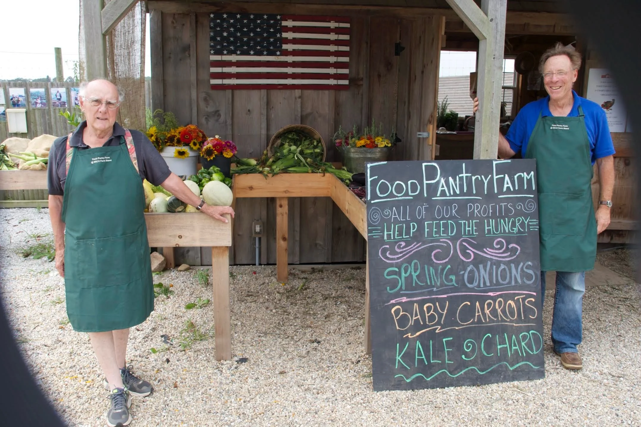 Two elderly men standing in front of a food pantry stall decorated with an American flag, with a chalkboard sign listing fresh vegetables including spring onions, baby carrots, kale, and chard. Both men are wearing green aprons and smiling.