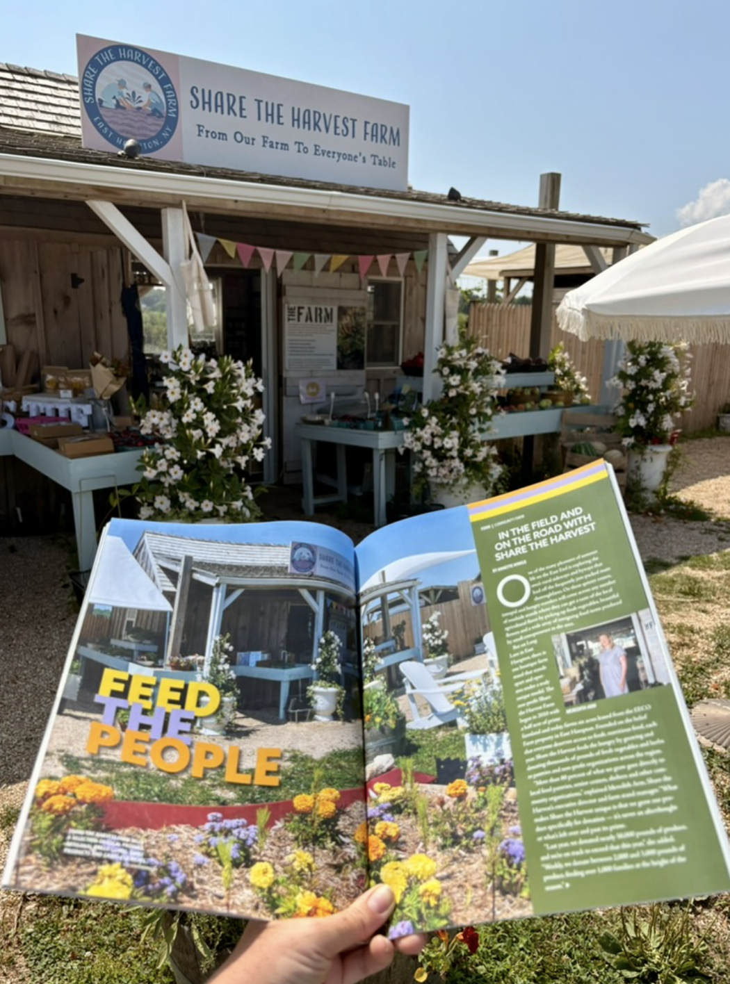A person holding an open magazine in front of a farm stand with a sign reading 'Share the Harvest Farm'. The farm stand has flowers and produce displays, and a small building with a sign about the farm. The magazine page features images of the farm and text encouraging feeding the people.