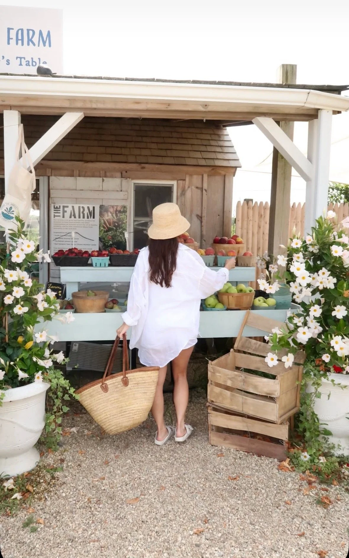 A woman wearing a white dress and a straw hat shopping for apples at an outdoor farm stand surrounded by flowers and wooden crates.
