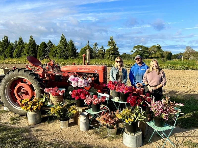 Three people standing in a field beside an antique red tractor decorated with pink and red flowers in pots and baskets, with additional flower arrangements on tables and chairs in front.