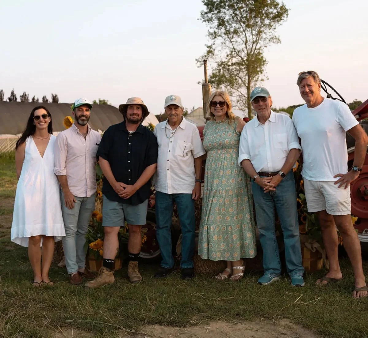 Group of eight people standing outdoors during sunset, smiling for a photo, with farm equipment and sunflowers in the background.