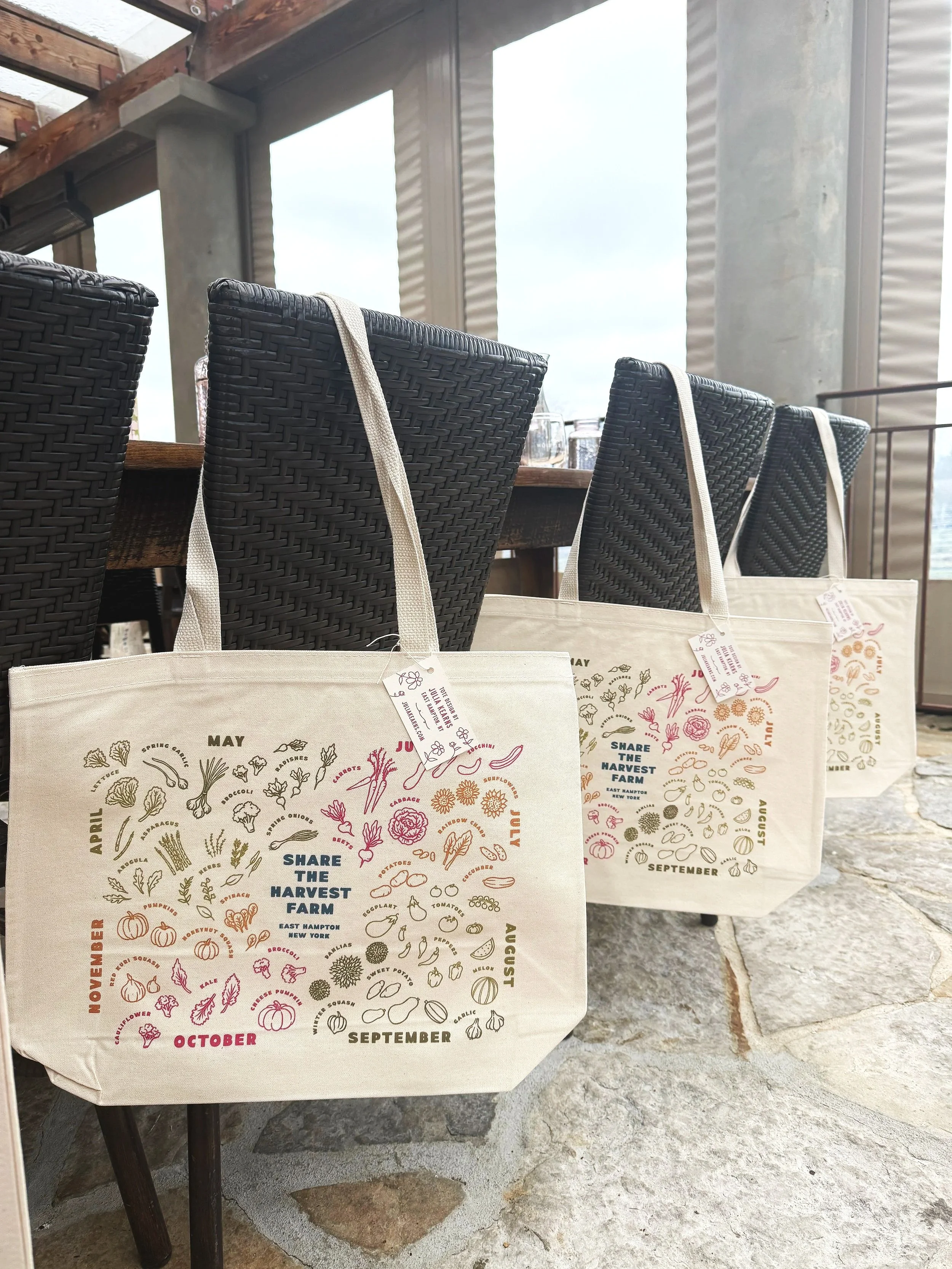 A row of tote bags hanging from a table with a rustic and industrial setting, including wooden beams and concrete pillars in the background. The bags feature colorful calendar illustrations for different months, with the phrase "Share the Harvest Farm" and various vegetable illustrations.