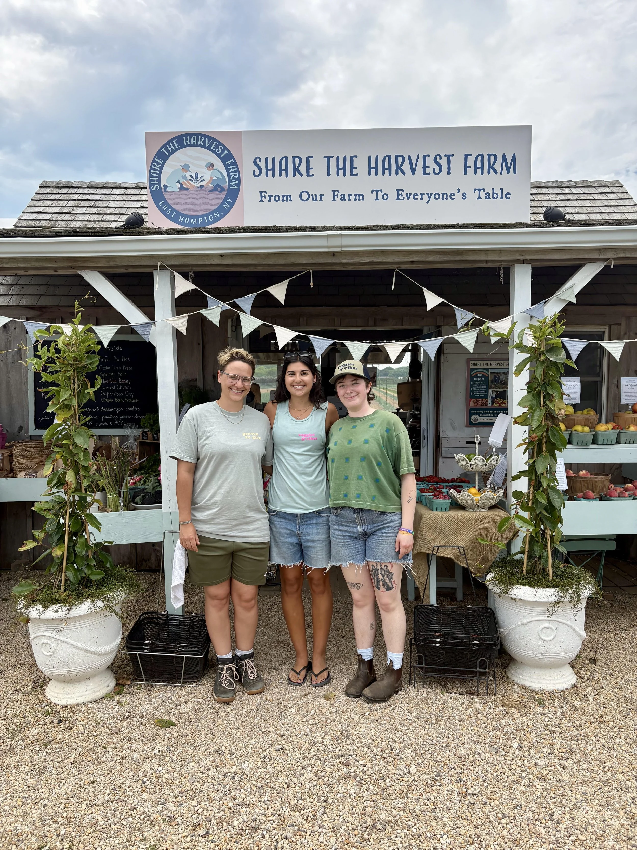 Three women standing in front of a farm stand with fruit, vegetables, and plants, smiling, and posing for the photo.