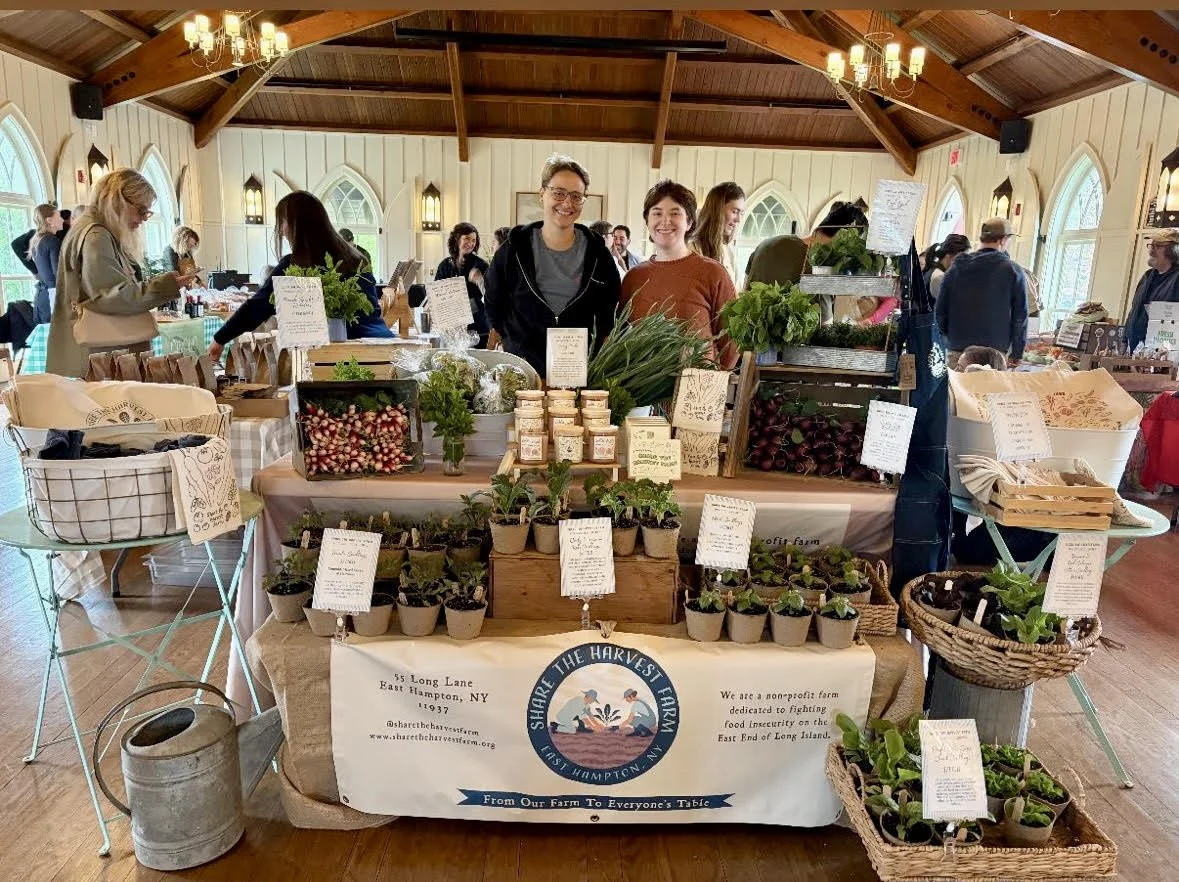 A farm booth at an indoor market displays herbs, plants, jars, and baked goods. Two women stand behind the table smiling. People browse other stalls in the background inside a wooden hall with arched windows and hanging lights.