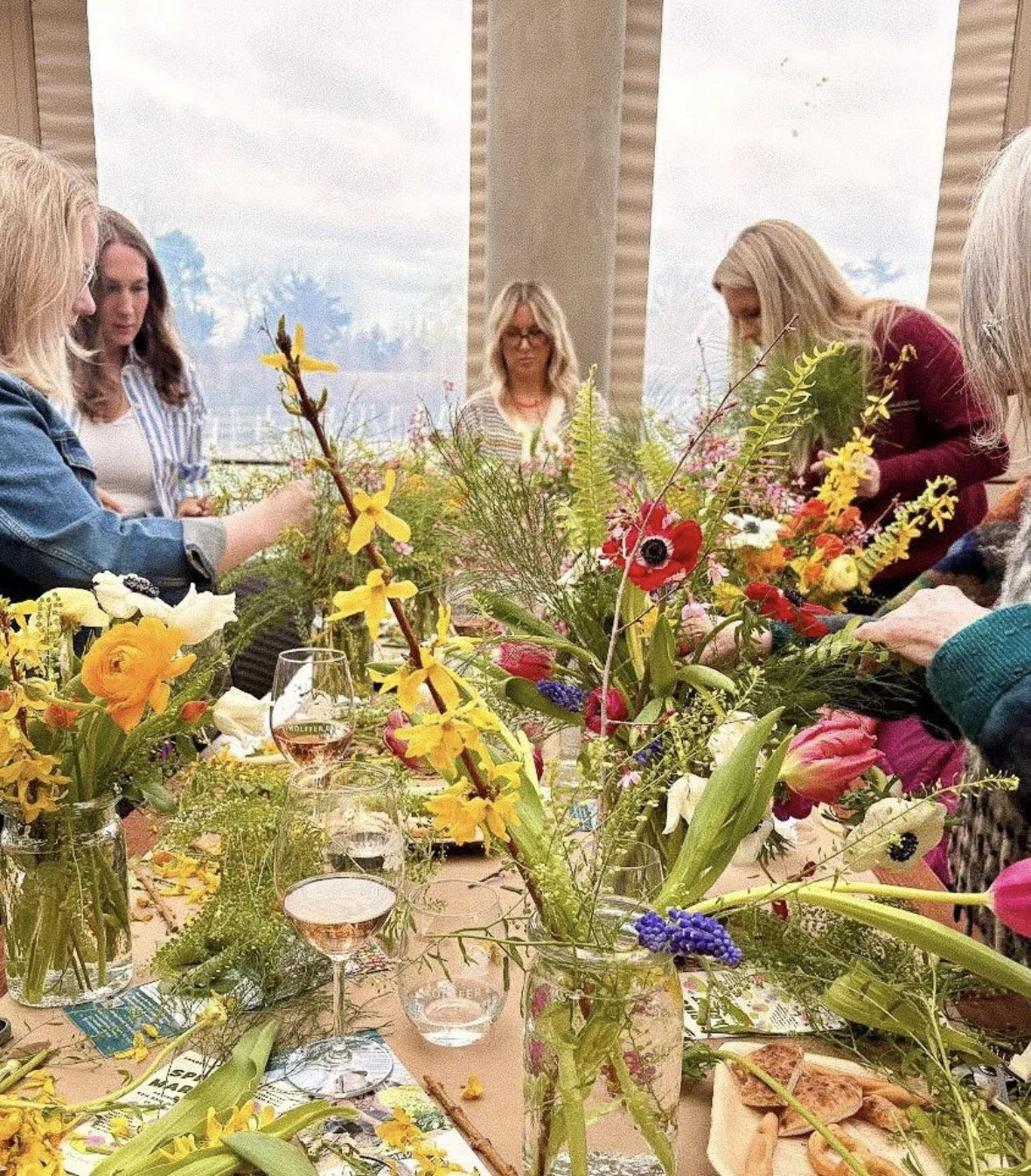 A group of women gather around a table filled with colorful flowers and floral arrangements for a flower arranging class or workshop.