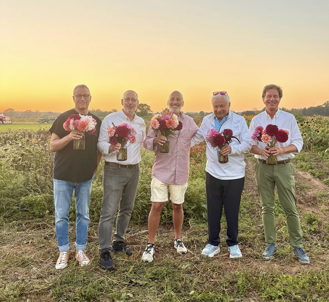 Five men standing in a field holding vases with pink and red flowers during sunset.