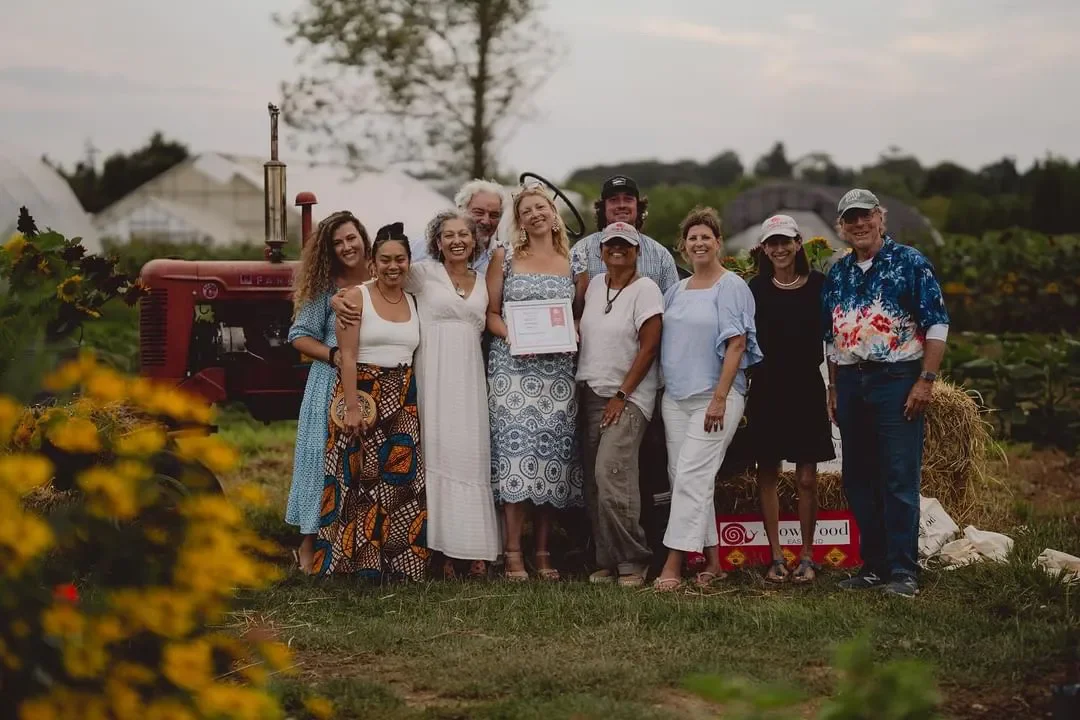 Group of ten people standing together outdoors in a field, some holding a certificate, with a tractor in the background, celebrating an achievement or event.