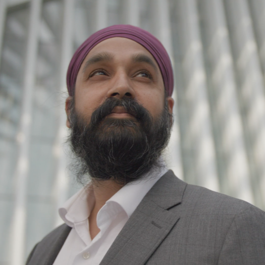 Portrait of bestselling author and religion scholar Simran Jeet Singh, a Sikh man wearing a purple turban with a dark beard, dressed in a gray suit and white shirt, standing outdoors with a modern building in the background.