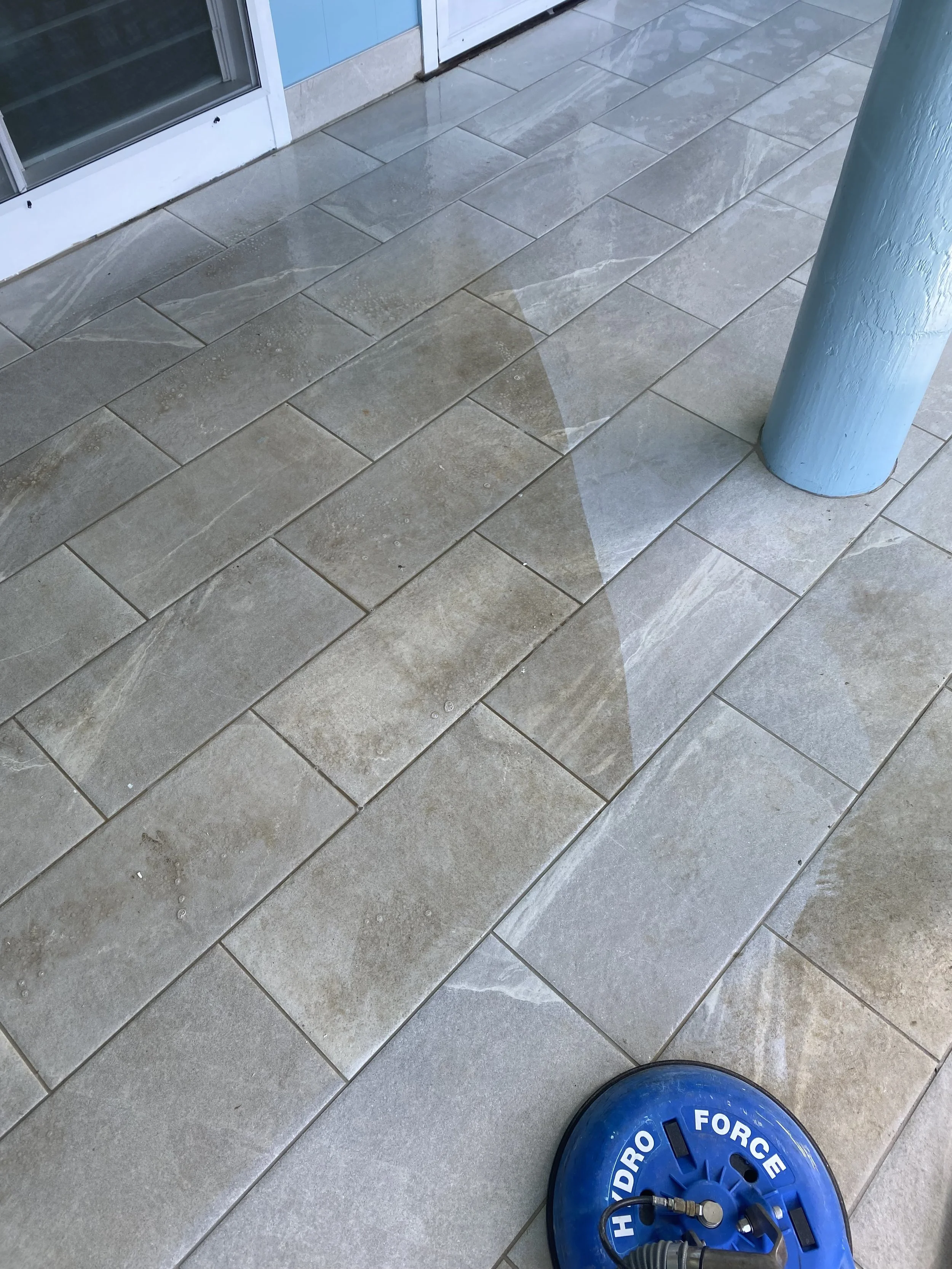 A tiled floor with a water puddle reflecting part of a door and a blue support pole, and a blue Hydro-FORCE pressure washer nozzle resting on the ground.