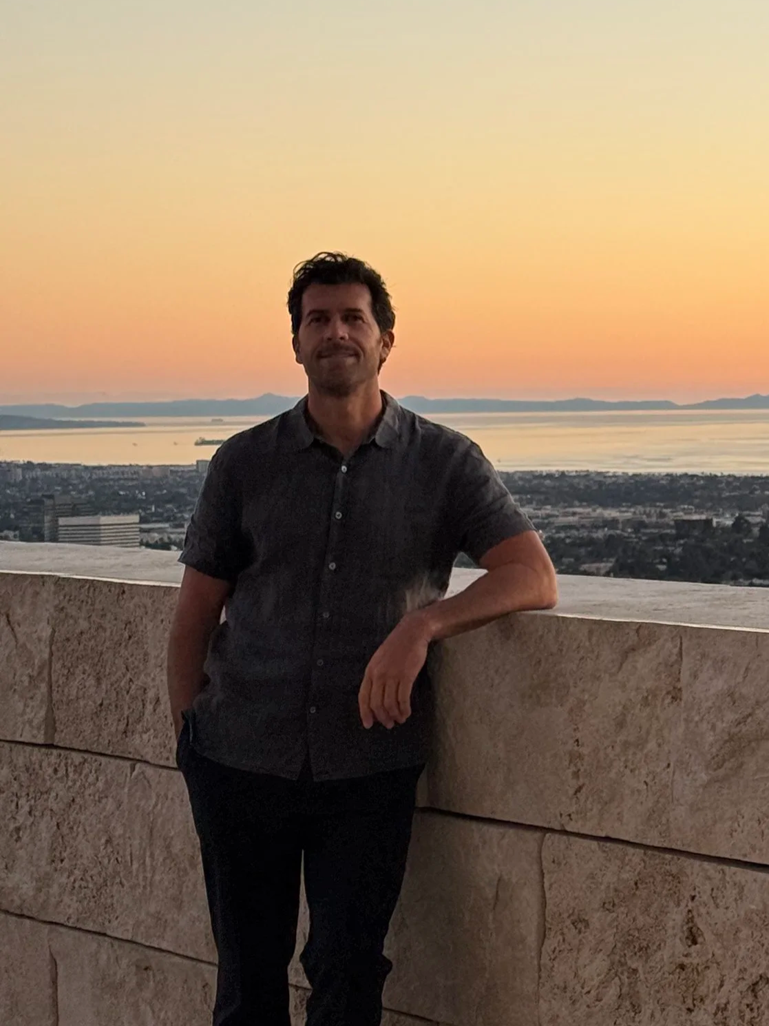 A man standing near a stone railing with a sunset over a body of water and distant mountains in the background. Kauai sunset.