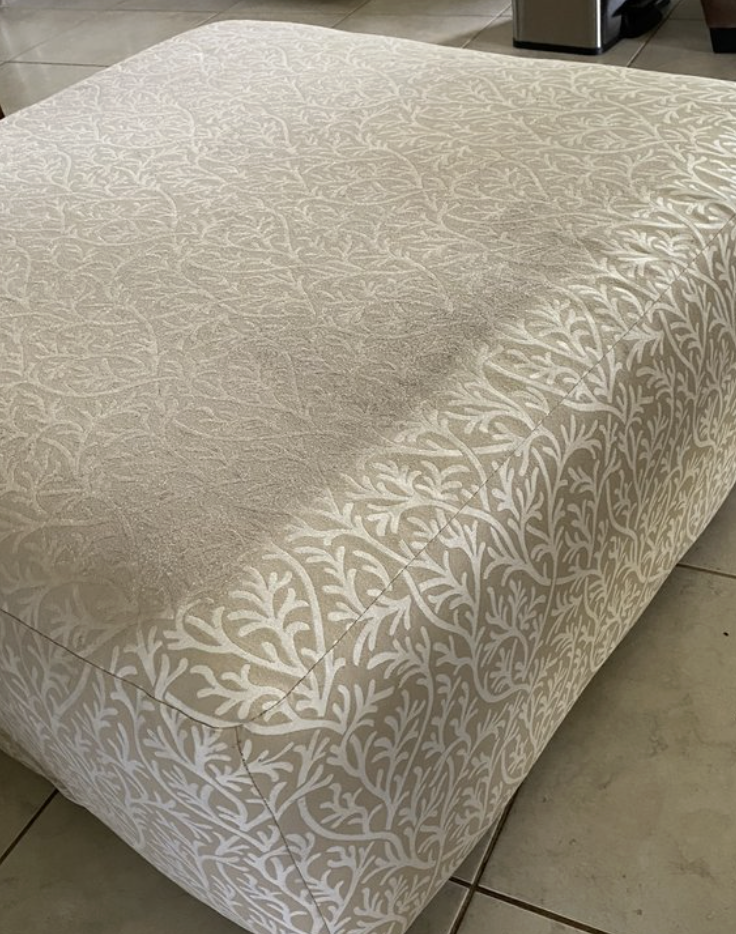Close-up of a beige upholstered ottoman with white leaf and vine pattern fabric, situated on a tiled floor.