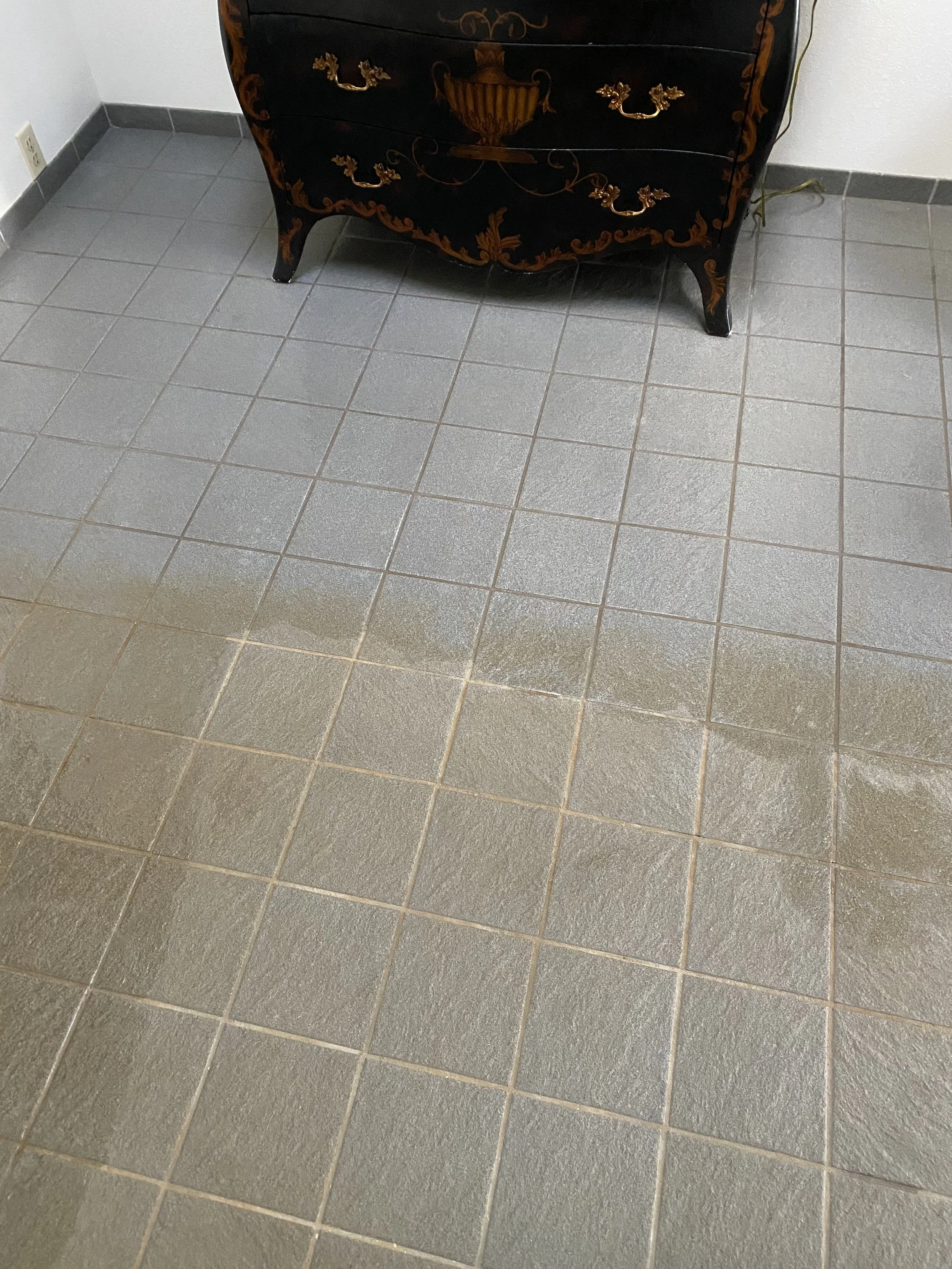 Two sections of tiled floor, one section appears clean and the other section appears dirty or wet, with a black dresser with gold accents against a white wall nearby.