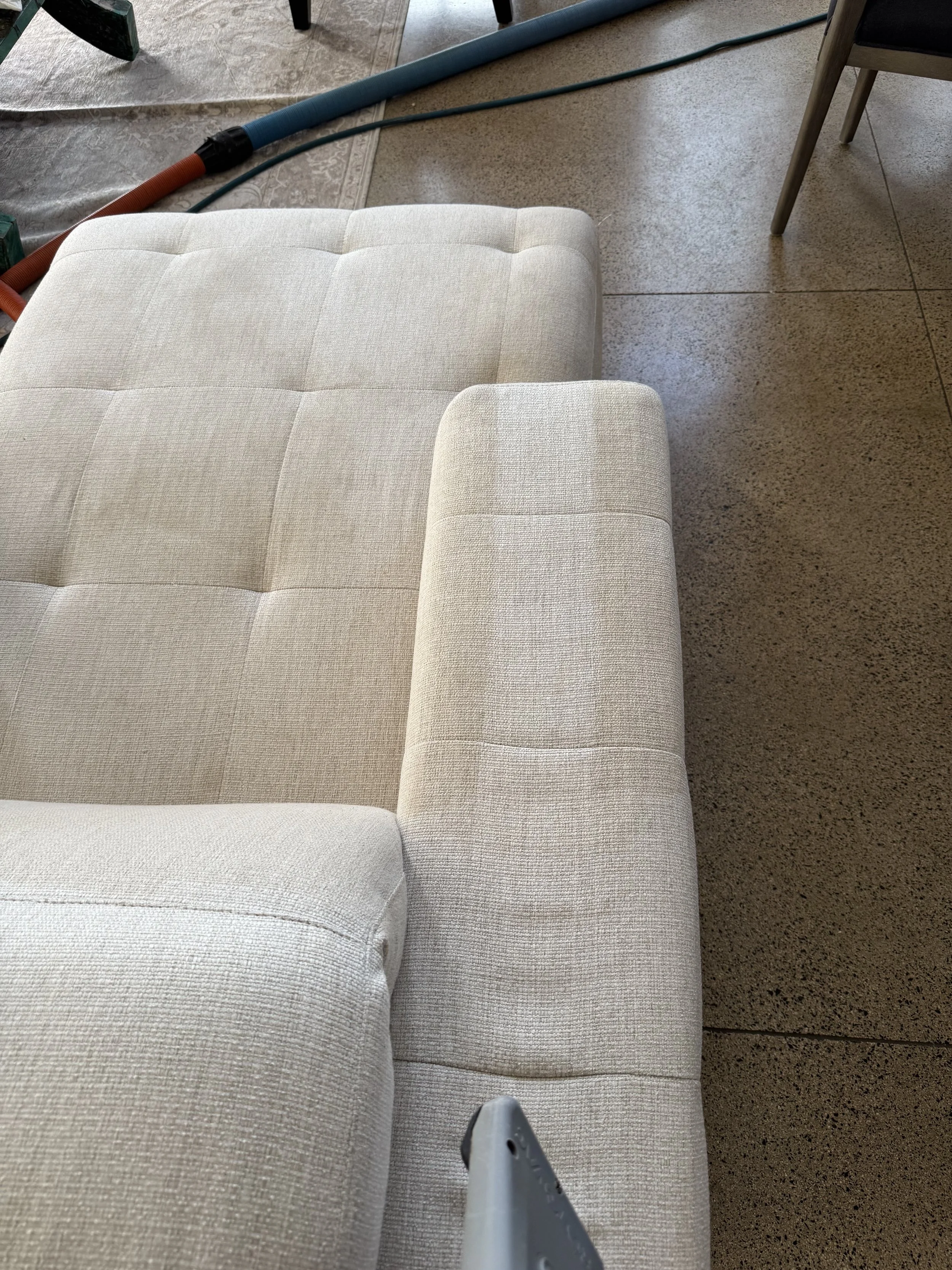 Close-up of a beige upholstered sofa with tufted cushions and a matching armrest in a room with terrazzo flooring, with some visible tools, hoses, and a chair in the background.
