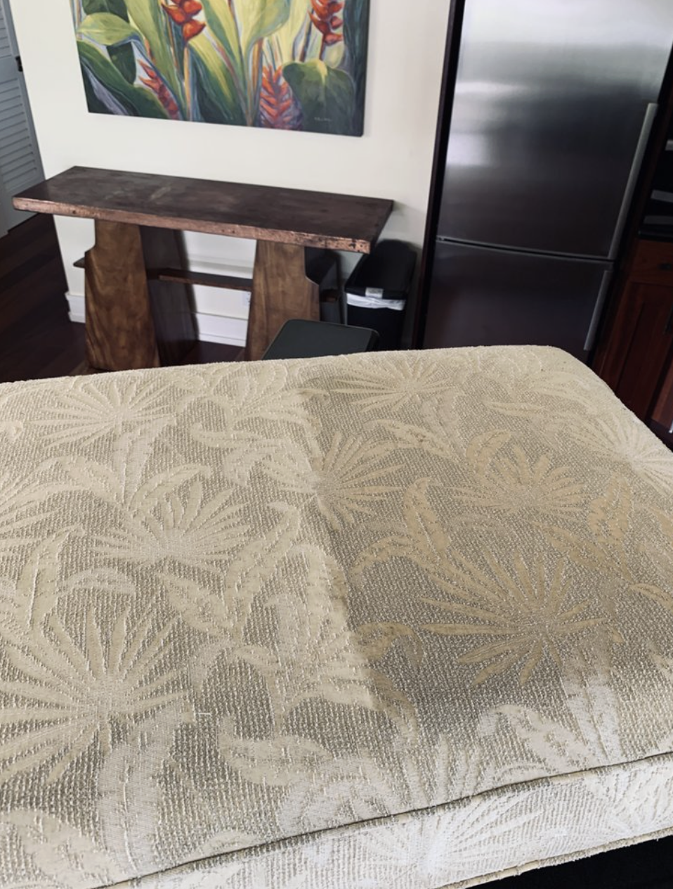 Part of a beige patterned fabric surface in the foreground, a wooden table against a wall with a colorful abstract painting, a black chair, a stainless steel refrigerator, and a small wooden cabinet in the background.