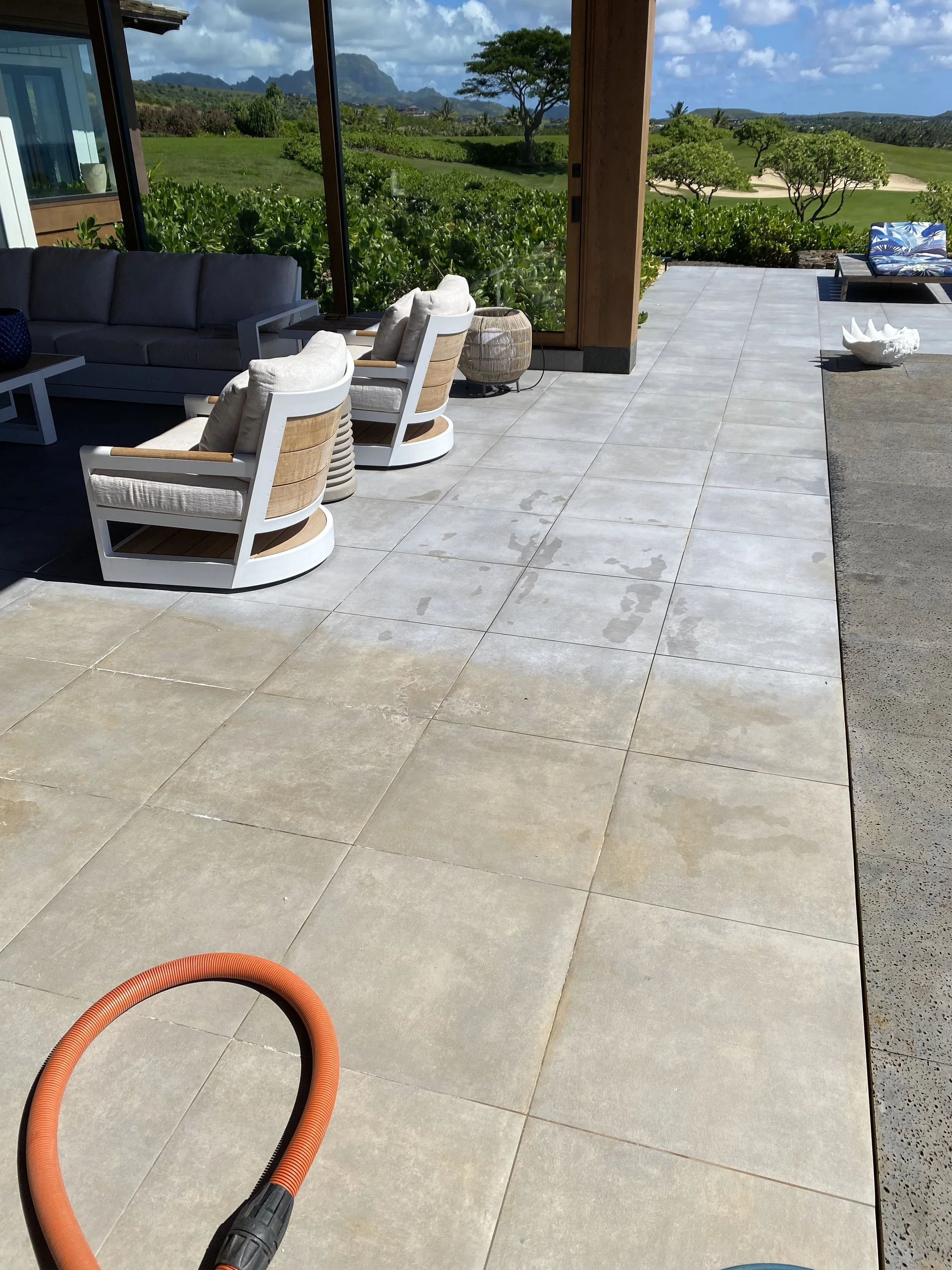 This image shows an outdoor patio with light-colored tiles, some wet from a recent cleaning or rain. There is an orange hose on the patio floor, and in the background, glass sliding doors reveal a lush green landscape with mountains and trees under a