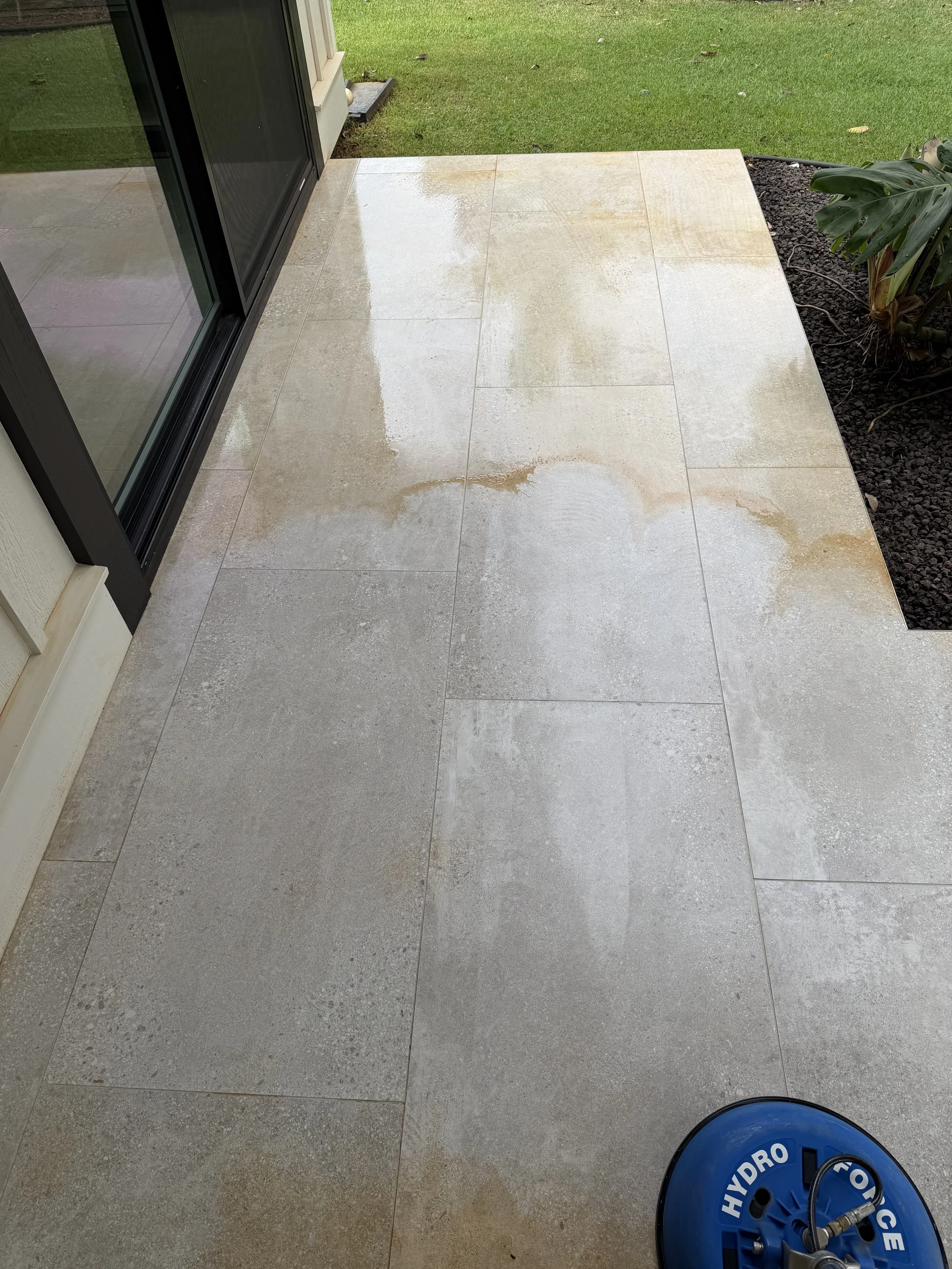 A wet, light-colored tiled patio with a sliding glass door on the left, a grassy lawn in the background, and a blue Hydro Septic toilet on the bottom right corner."