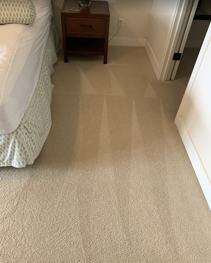 Clean beige carpet with visible vacuum lines in a bedroom next to a bed and a wooden nightstand.