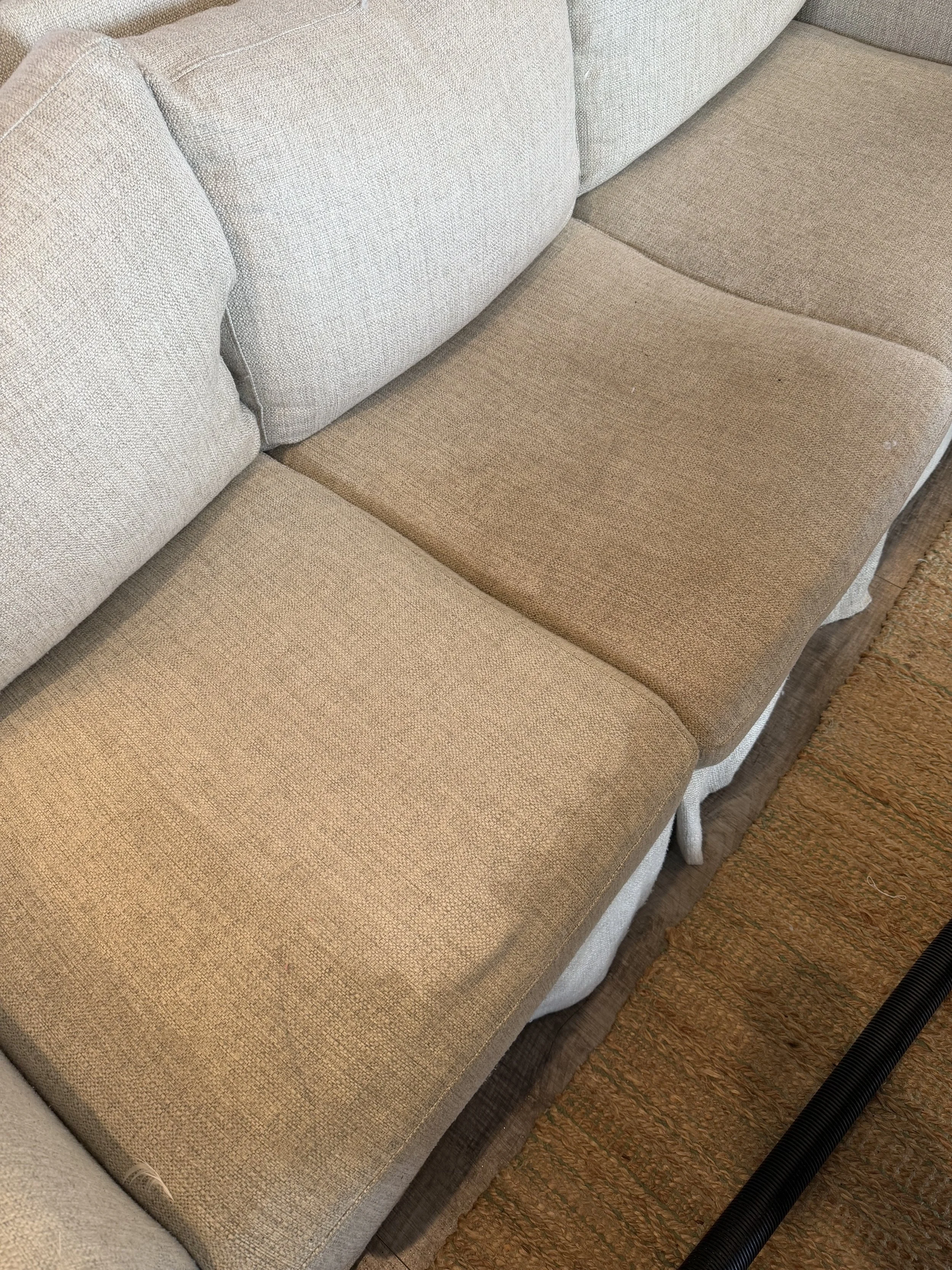 Close-up of a two-tone beige fabric sofa with light beige cushions and darker beige seat cushions, next to a textured brown rug and black vacuum hose.