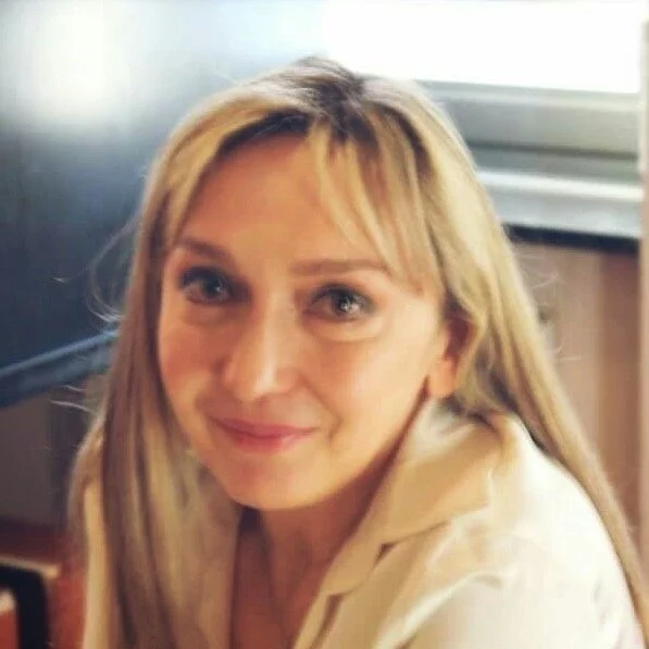 A woman with blonde hair and a light-colored top, sitting indoors near a window