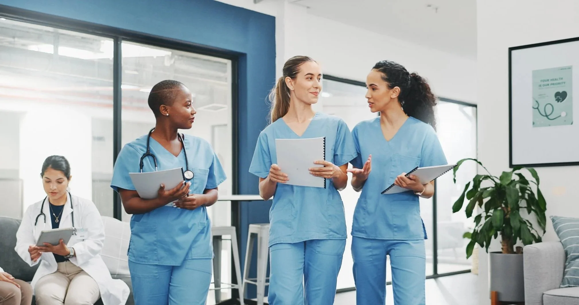 Four healthcare professionals in scrubs and a doctor in a white coat in a modern medical facility, engaged in a discussion, with a woman holding a tablet seated on a couch in the background.