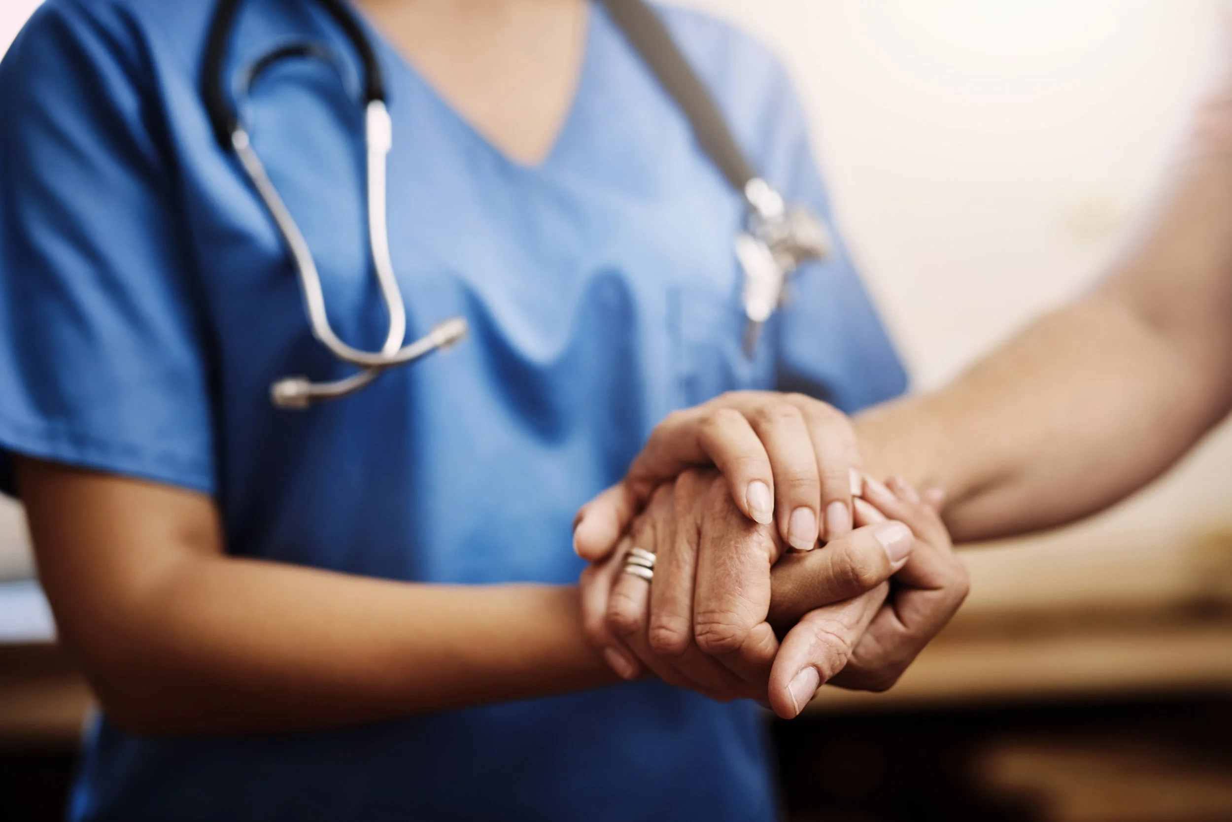 A healthcare professional in a blue uniform with a stethoscope around their neck is holding an elderly person's hand, showing care and compassion.