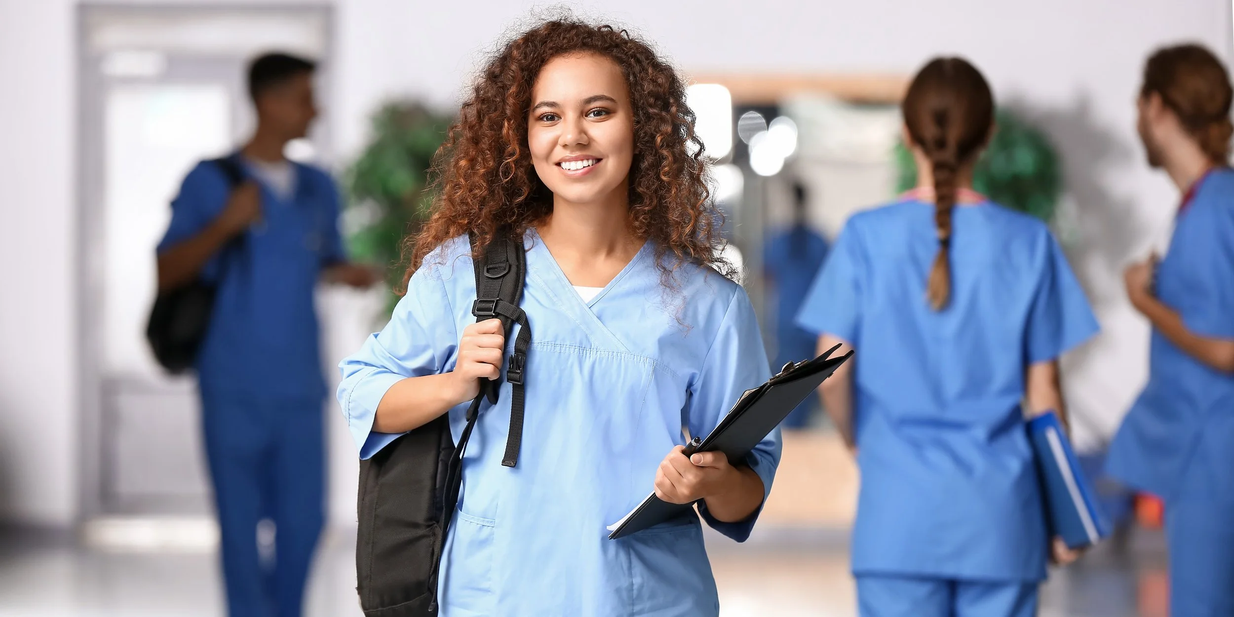 A young woman in blue scrubs smiling while holding a clipboard and backpack, with other people in similar scrubs in the background.