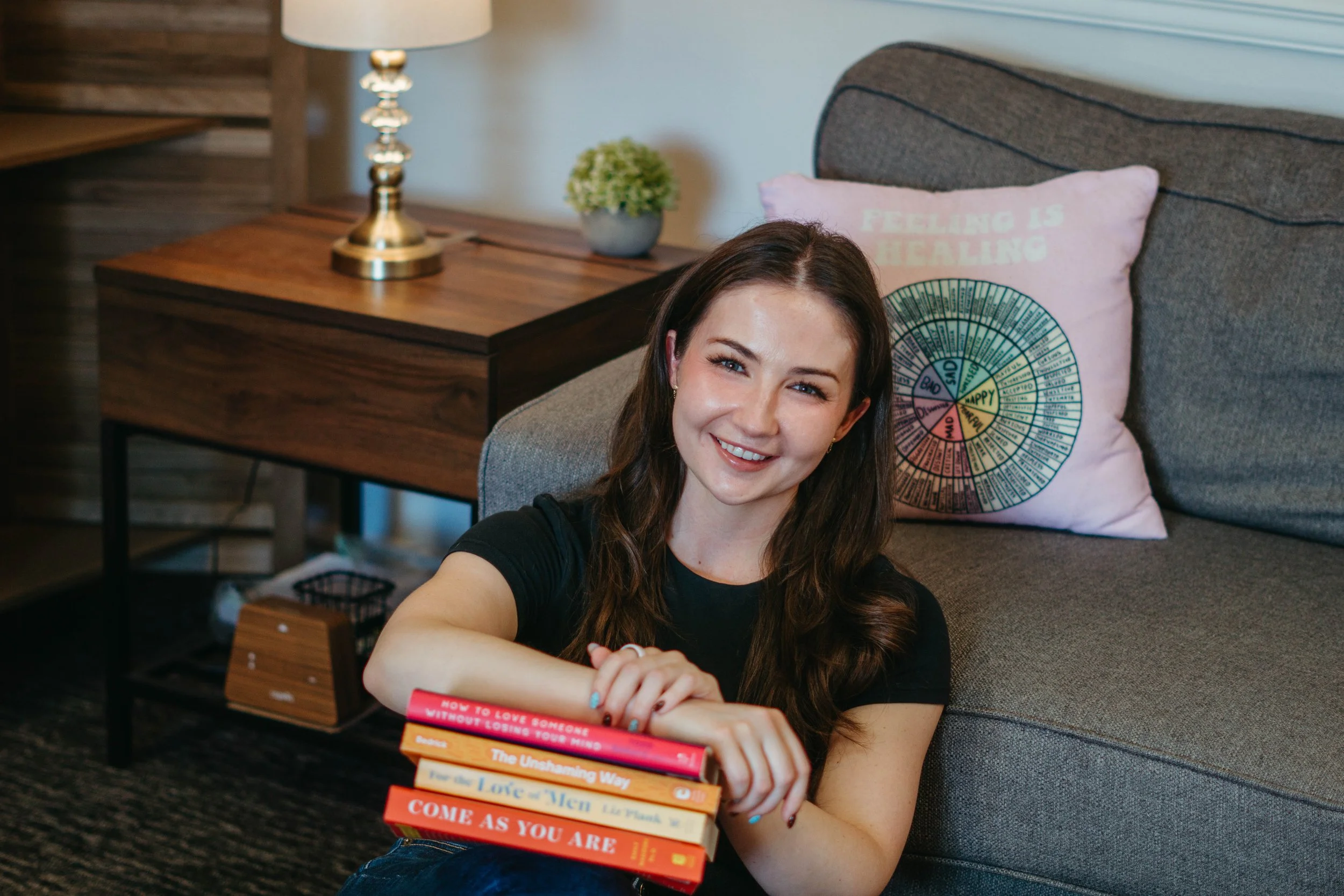 A woman sitting on the floor in front of a gray couch, smiling at the camera, holding a stack of books with colorful spines, with a pink pillow with a circular chart behind her and a wooden side table with a lamp and a small potted plant in the background.
