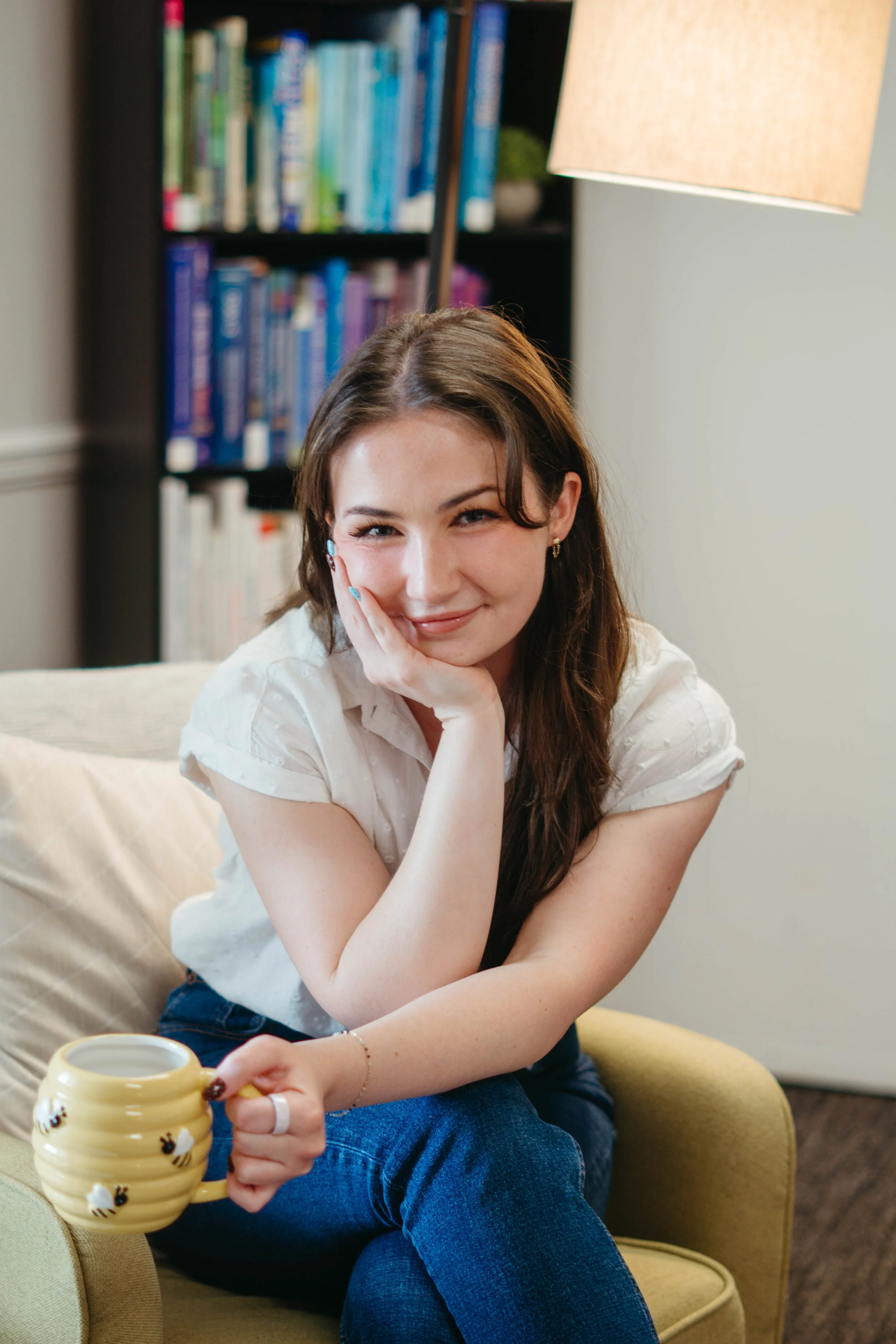 A young woman sitting on a yellow armchair, holding a yellow bee-themed mug, with a smile, in a cozy room with a bookshelf behind her and a beige lampshade overhead.