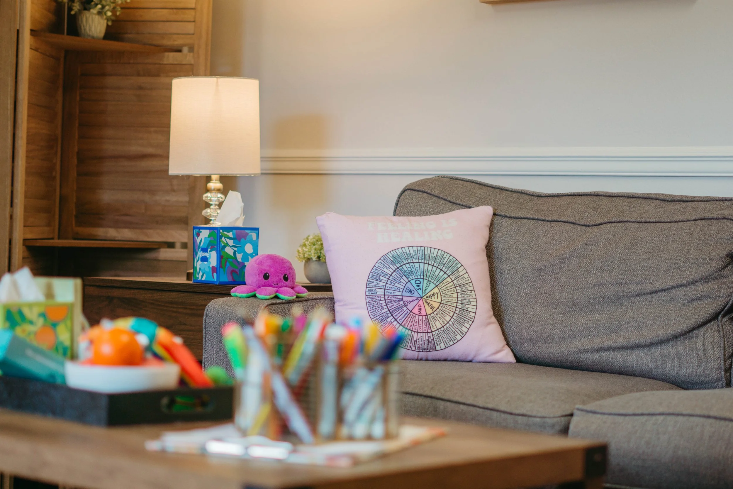 A cozy living room corner with a beige couch adorned with pink and colorful cushions, including one with a wellness wheel. A wooden shelf with a potted plant and lamps sits behind. The foreground features colorful decorated cupcakes on a table.