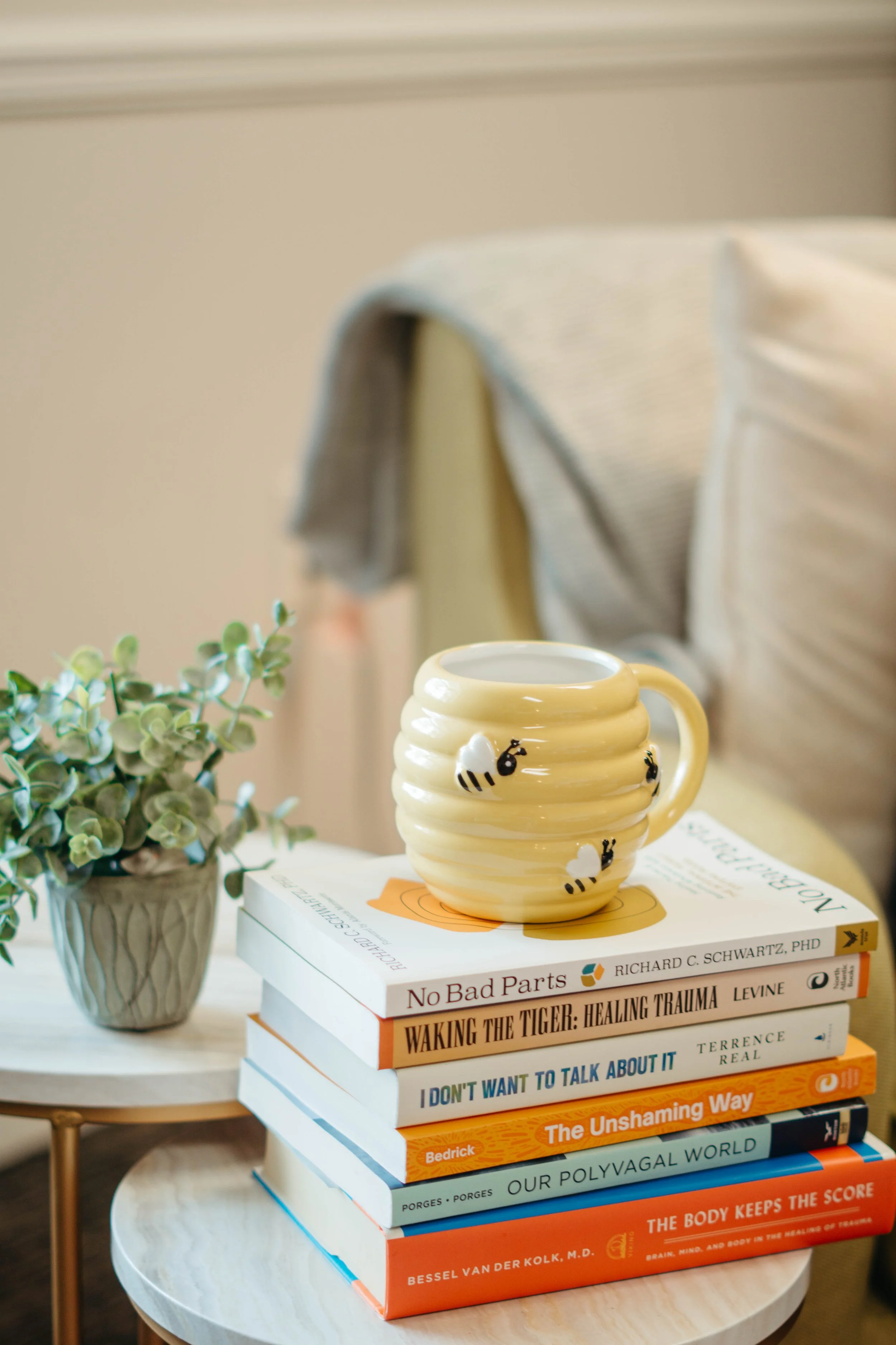 Stack of books with a beige mug decorated with bees on top, placed on a small round wooden table with a plant in a gray pot nearby.
