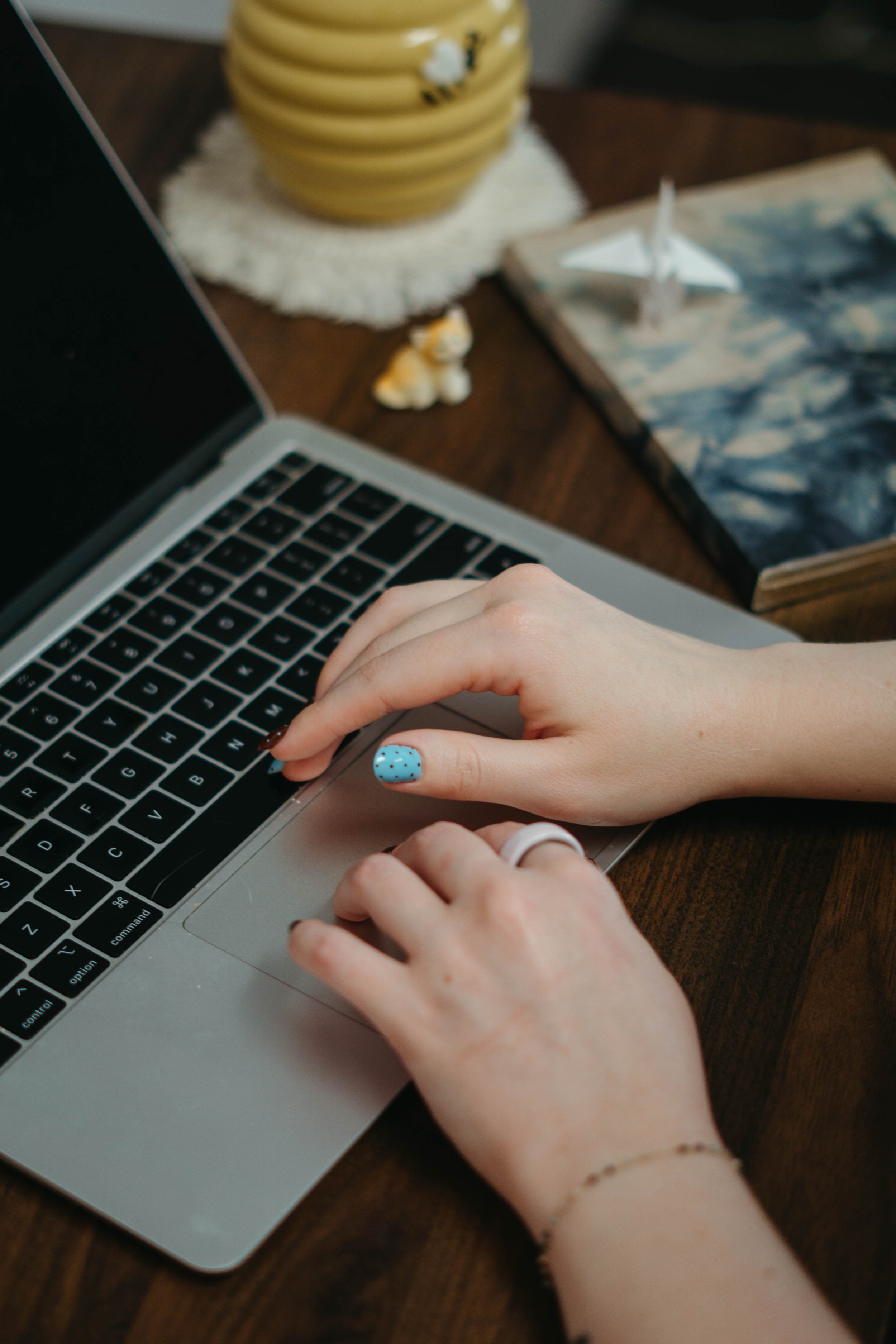 Person using a laptop with a yellow striped lampshade, a small ceramic dog figurine, a notebook with landscape photo, and a book with a sailboat on the cover on a wooden table.