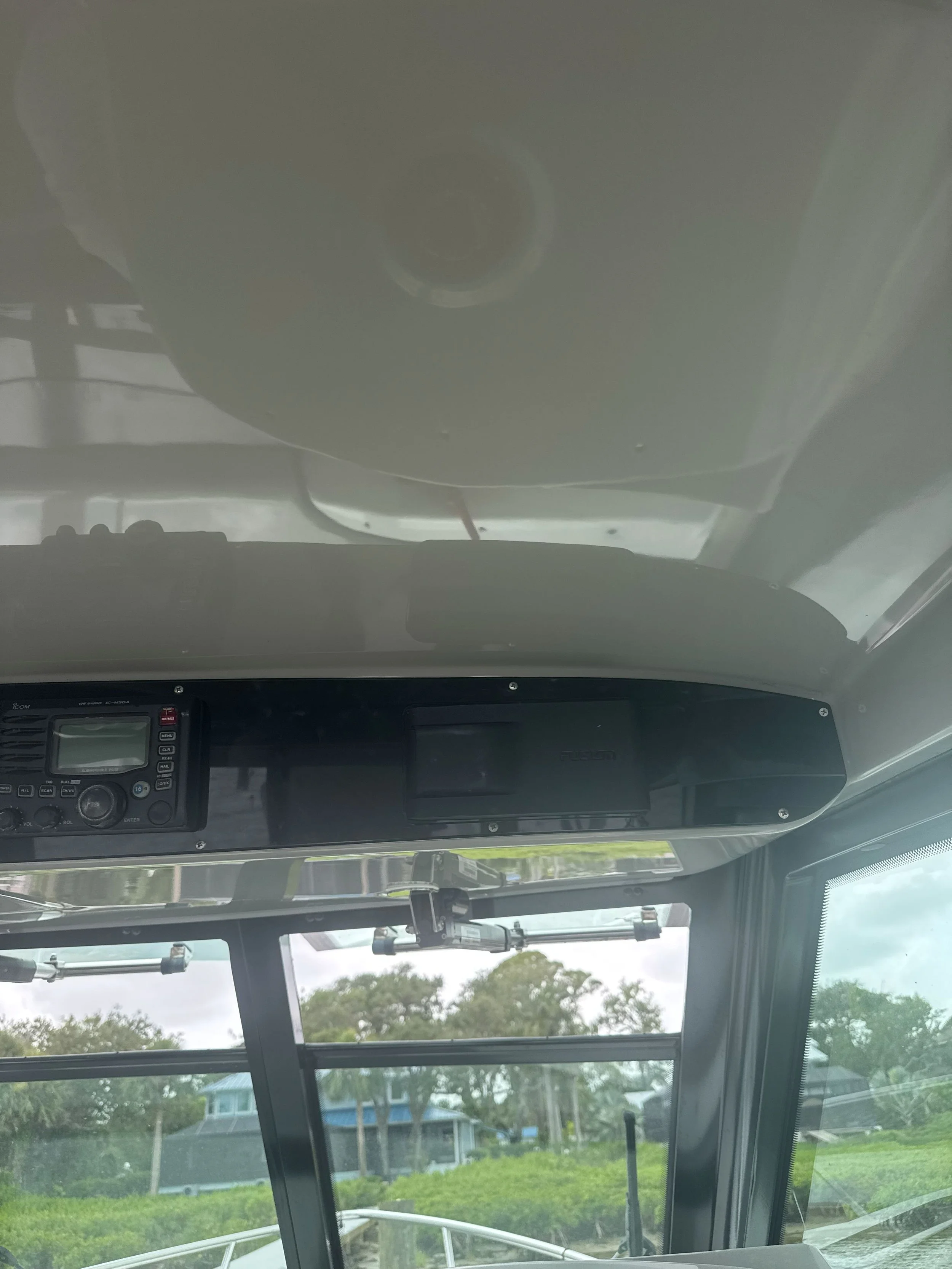 Interior view of a boat pilot's cabin showing the navigation and communication equipment, windows with a view of trees and houses outside.