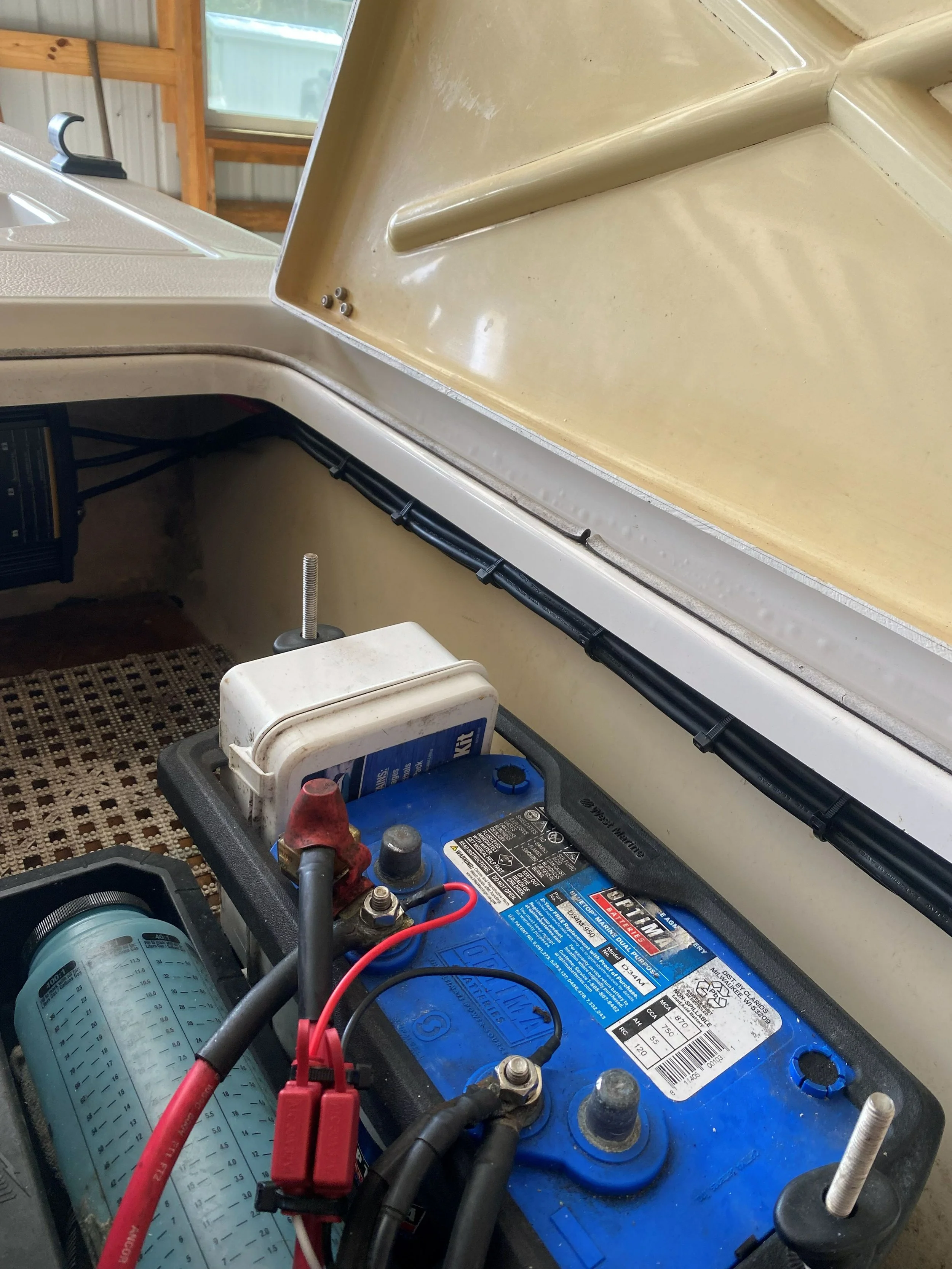 A boat battery connected with red and black cables, with a battery charger nearby, inside the boat's engine compartment with the cover open.