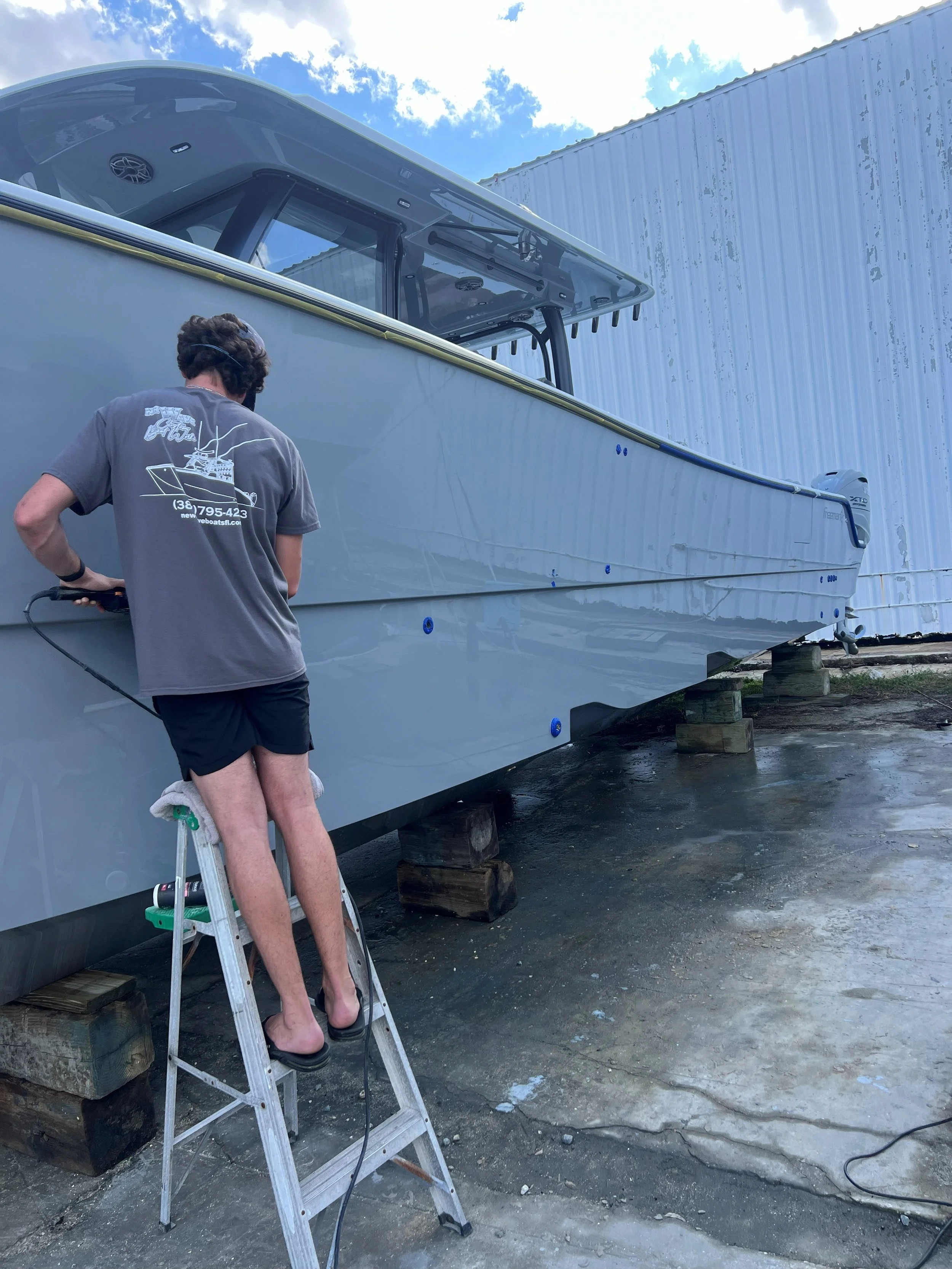 A man standing on a stepladder cleaning the hull of a large, white boat that is stored on wooden blocks outside near a metal building.