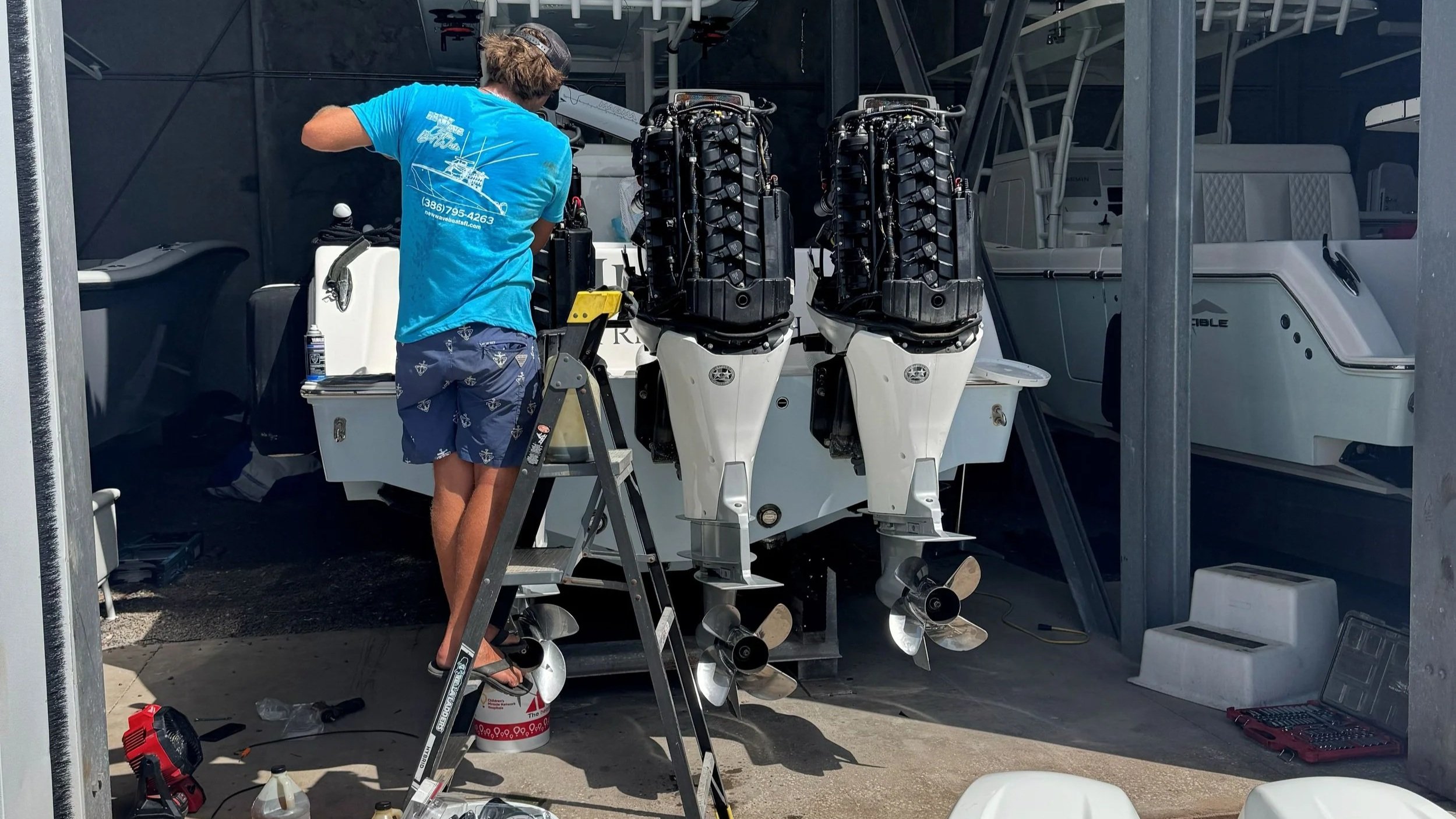A person working on a boat with dual engines, wearing a blue t-shirt and shorts, standing on a ladder in a garage or storage area with tools and equipment around.
