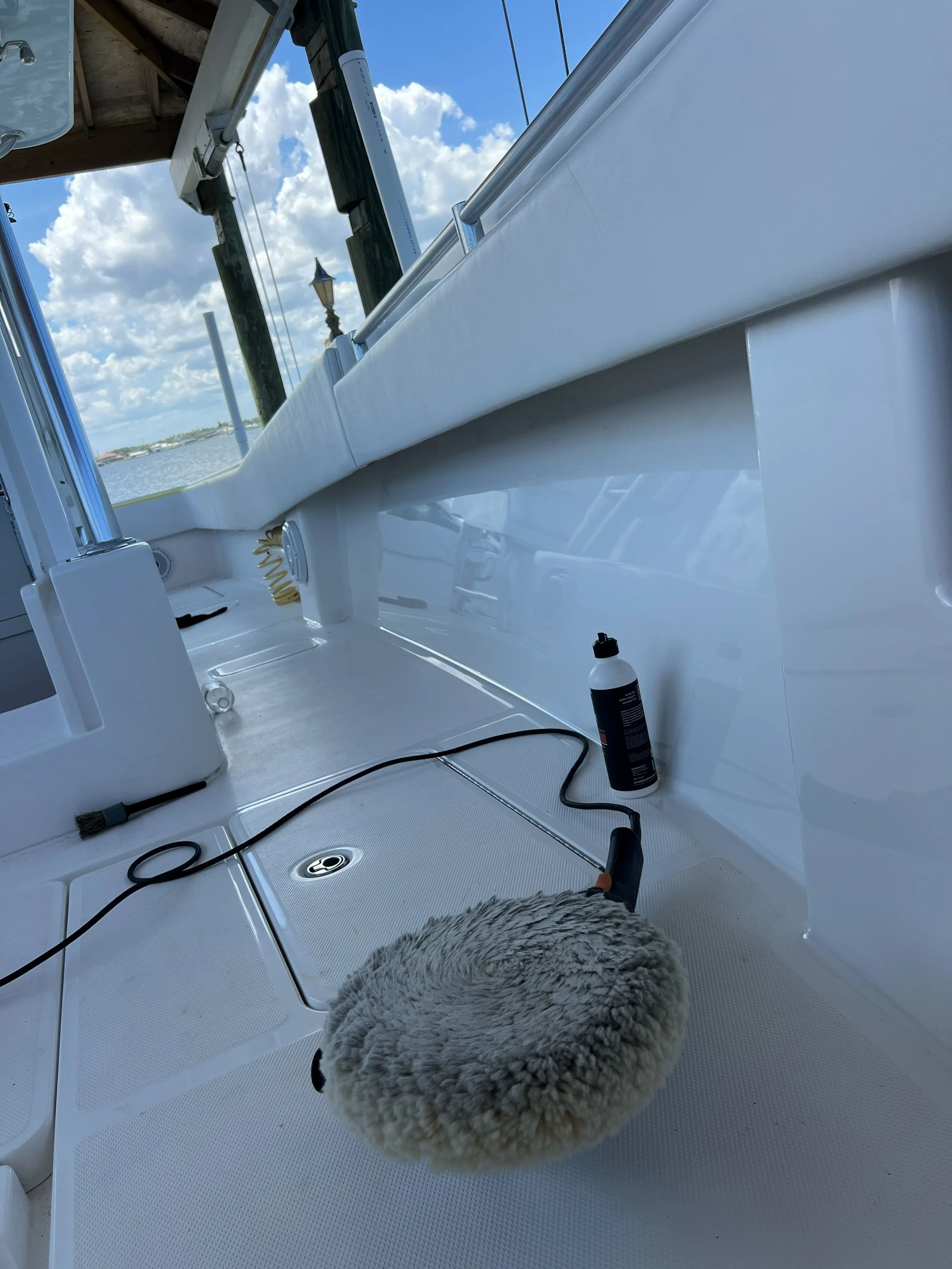 The image depicts the deck of a boat with cleaning equipment, including a gray cleaning pad and a bottle of cleaning solution. The boat is docked near a pier with a body of water visible in the background, under a sky with scattered clouds.