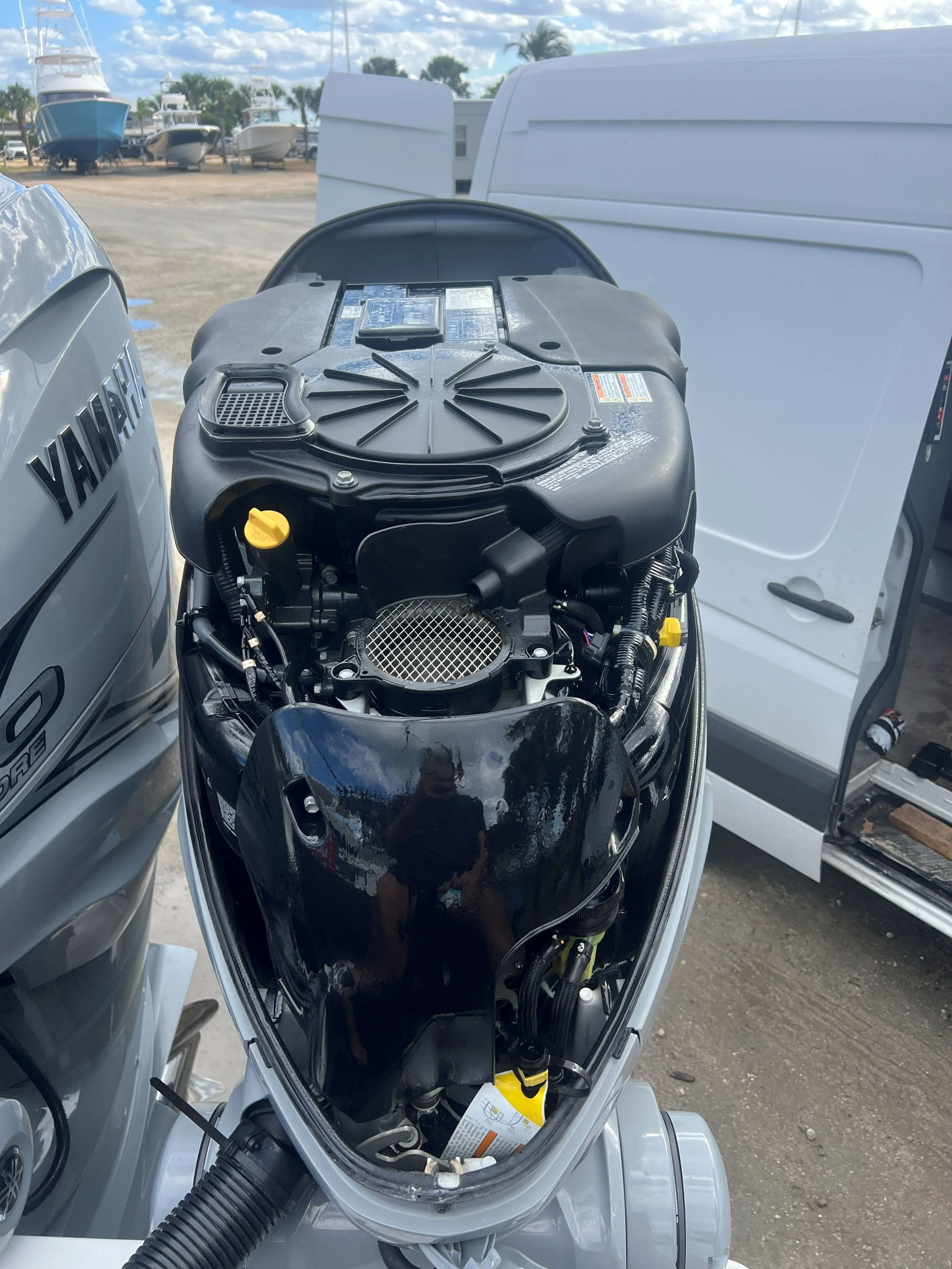 Close-up view of a boat's outboard motor with the cover removed, showing the engine and components, with boats in the background at a marina or boatyard.