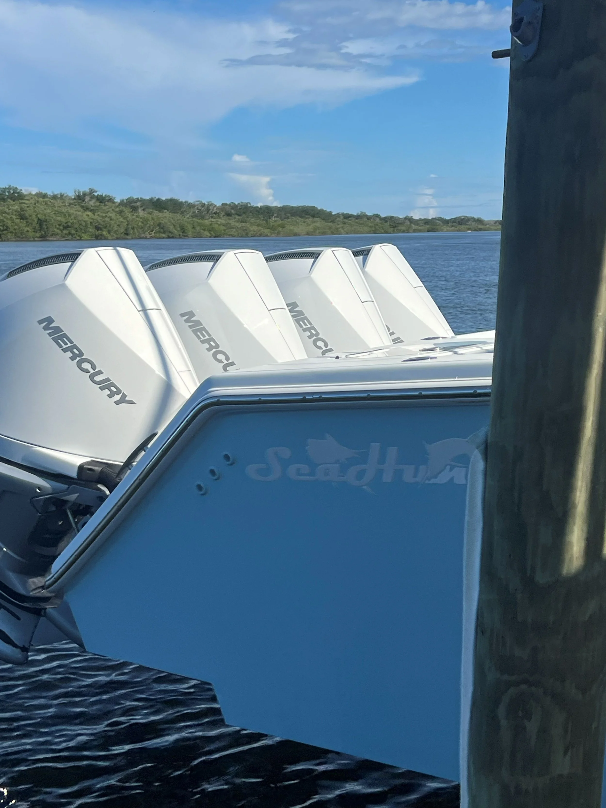 Four white Mercury outboard motors on a boat, with a blue sky, water, and green trees in the background.