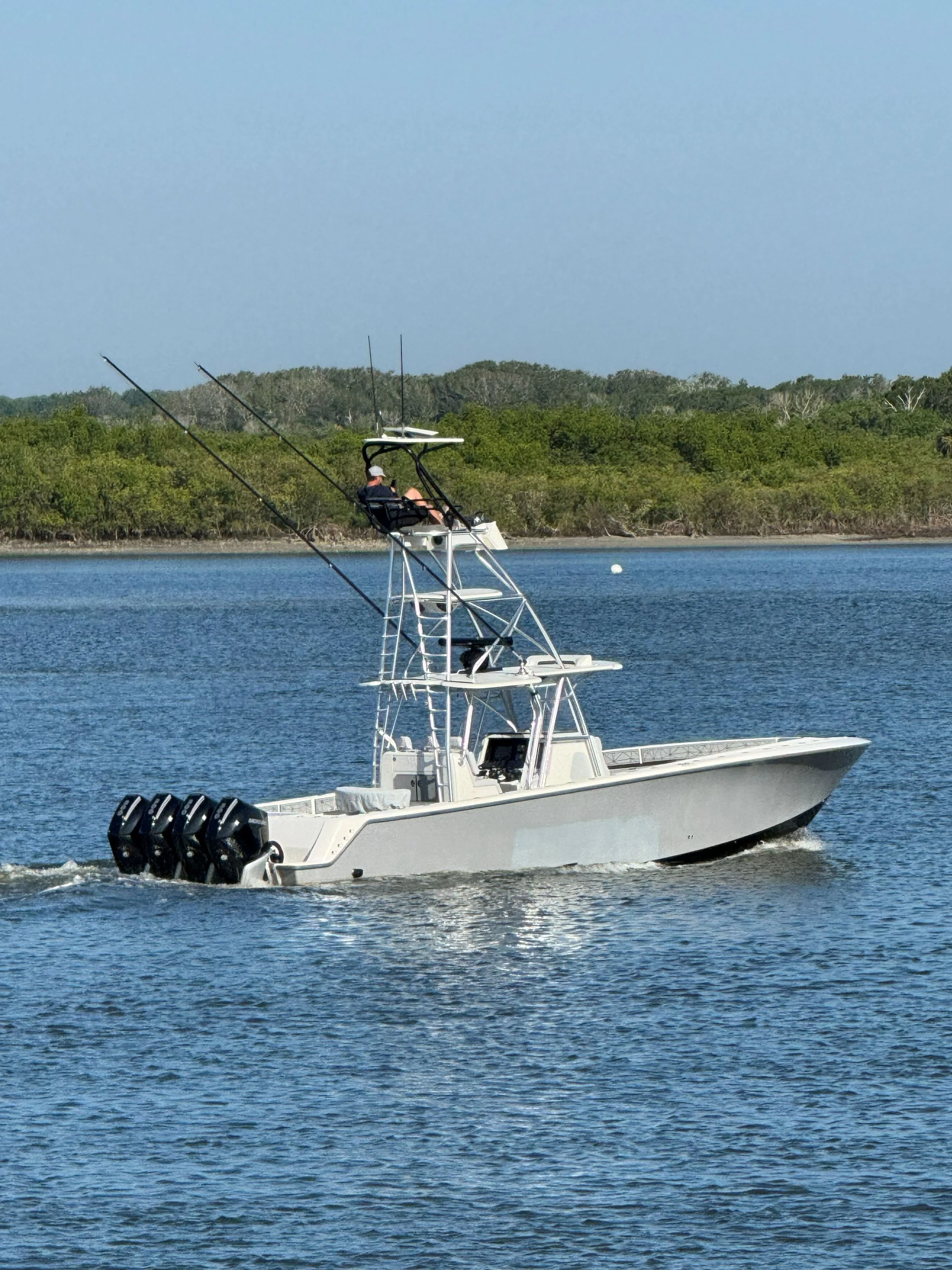 A white fishing boat with four outboard motors sailing on calm water, with a person sitting in the upper seating area, fishing rods mounted on the boat, and trees in the background under a blue sky.