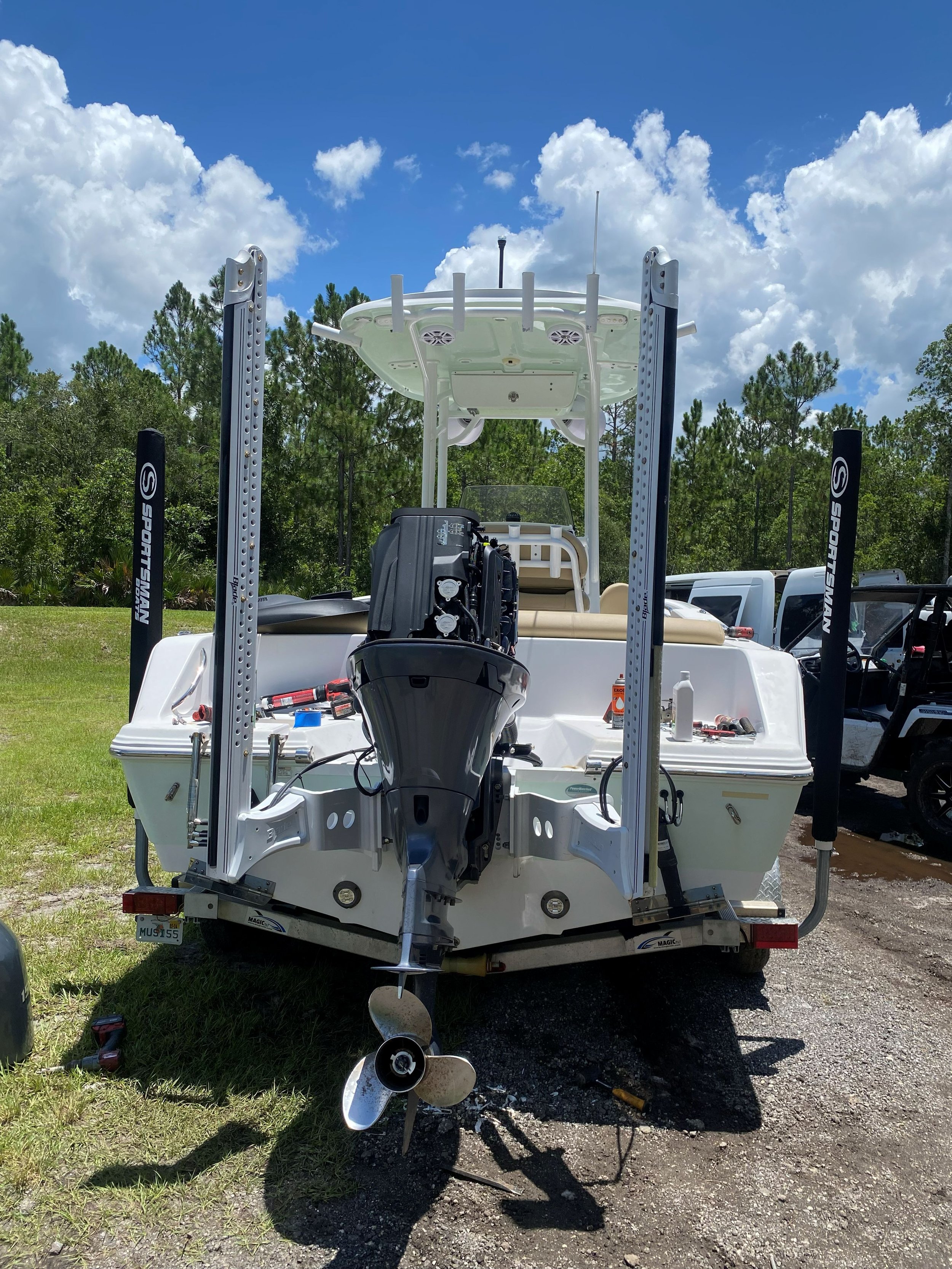 A white boat on a trailer with a black outboard motor, boat lift supports, and fishing equipment, parked on grass and gravel with trees and a partly cloudy sky in the background.