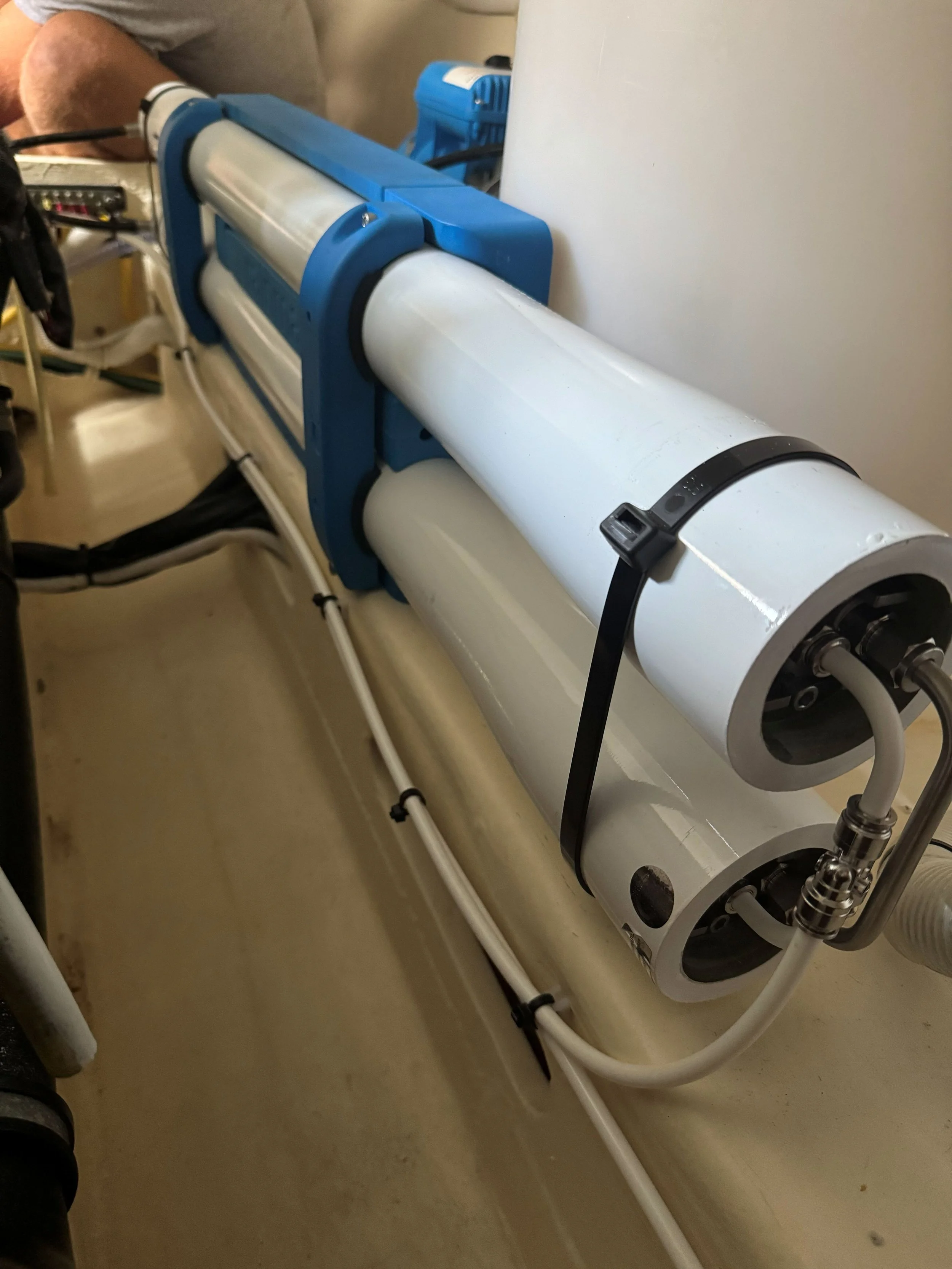 Close-up of a large scientific or industrial device with white cylindrical components, blue brackets, and connected tubes, mounted on a beige surface with wiring and cables nearby.