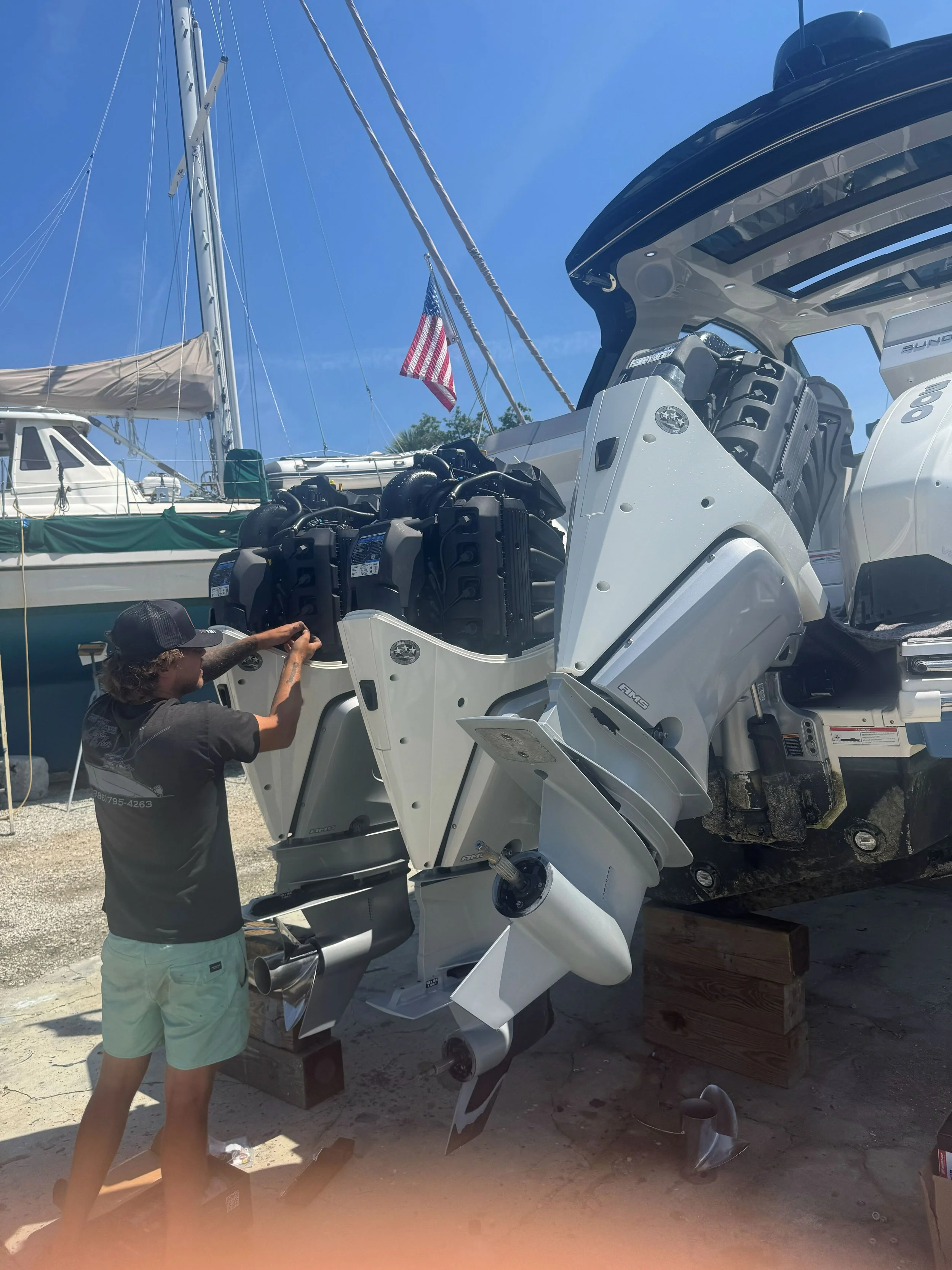 A person working on a large boat with multiple outboard engines, on land near a marina, with a sailboat and American flag visible in the background.
