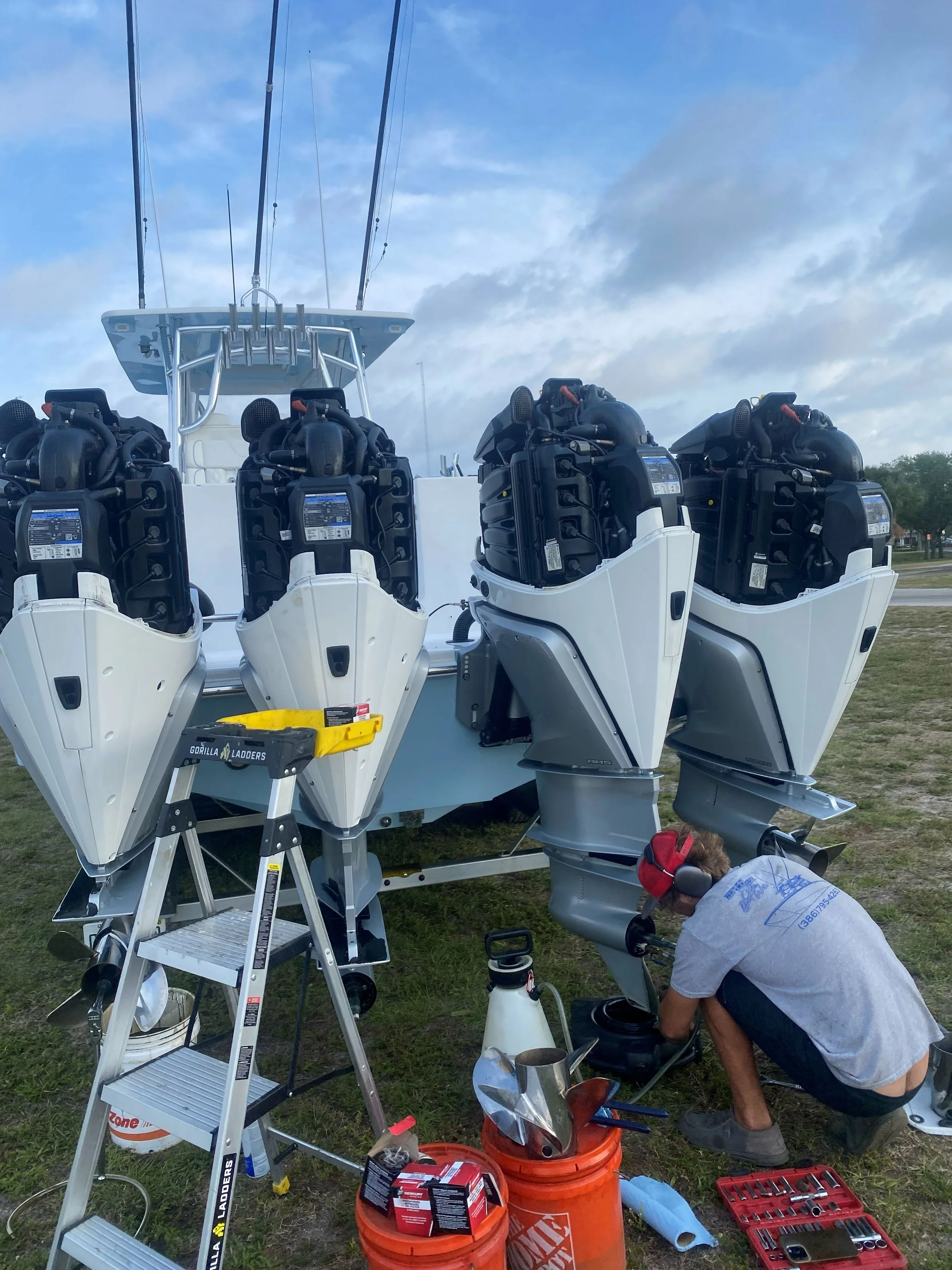 A person wearing a gray t-shirt, red ear protection, and headphones is working on a boat with four outboard motors. The person is kneeling and using tools, with ladders, containers, and toolboxes around.